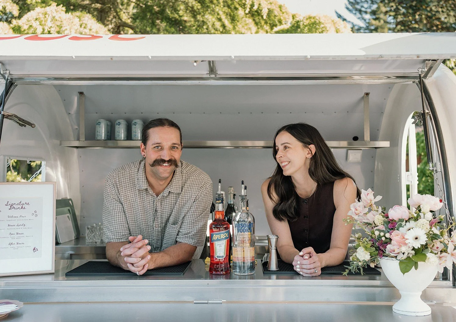A man and woman are standing behind a food truck counter, smiling. The man has a mustache and is wearing a checked shirt, while the woman has long dark hair and is wearing a black sleeveless top. There are bottles of alcohol, a mixer, and a flower arrangement on the counter, with trees in the background.