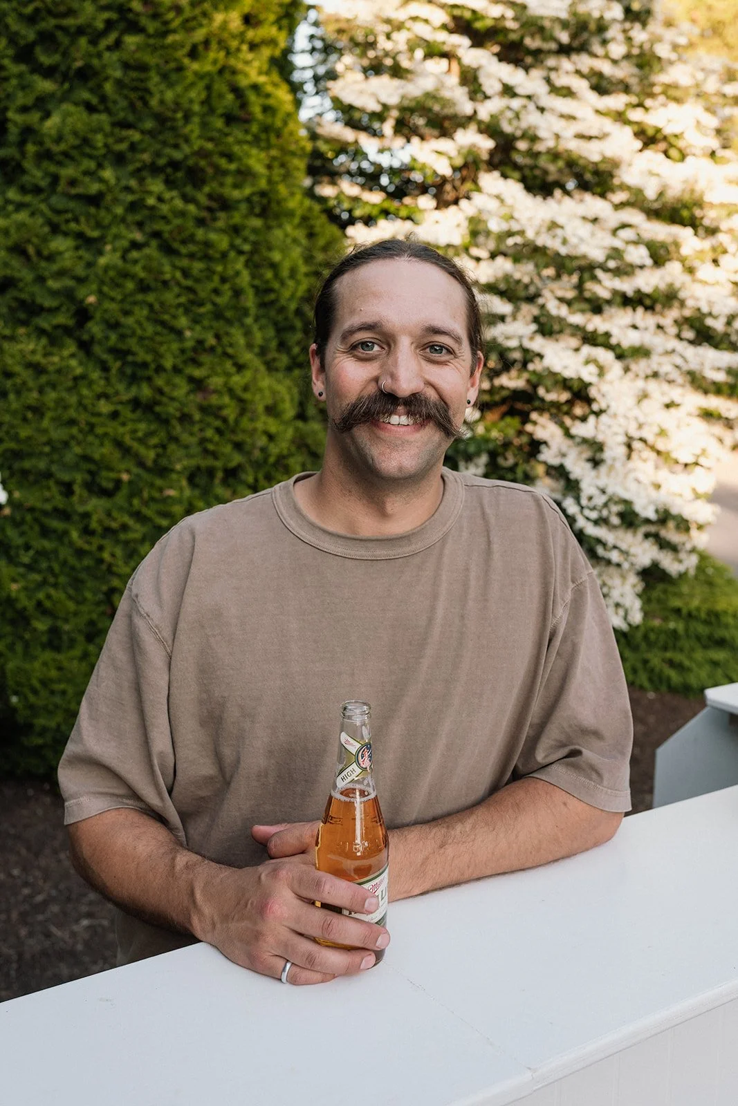 A man with a mustache, wearing a beige t-shirt, holding a bottle of beer, smiling, outdoors in a garden with green bushes and a flowering tree in the background.