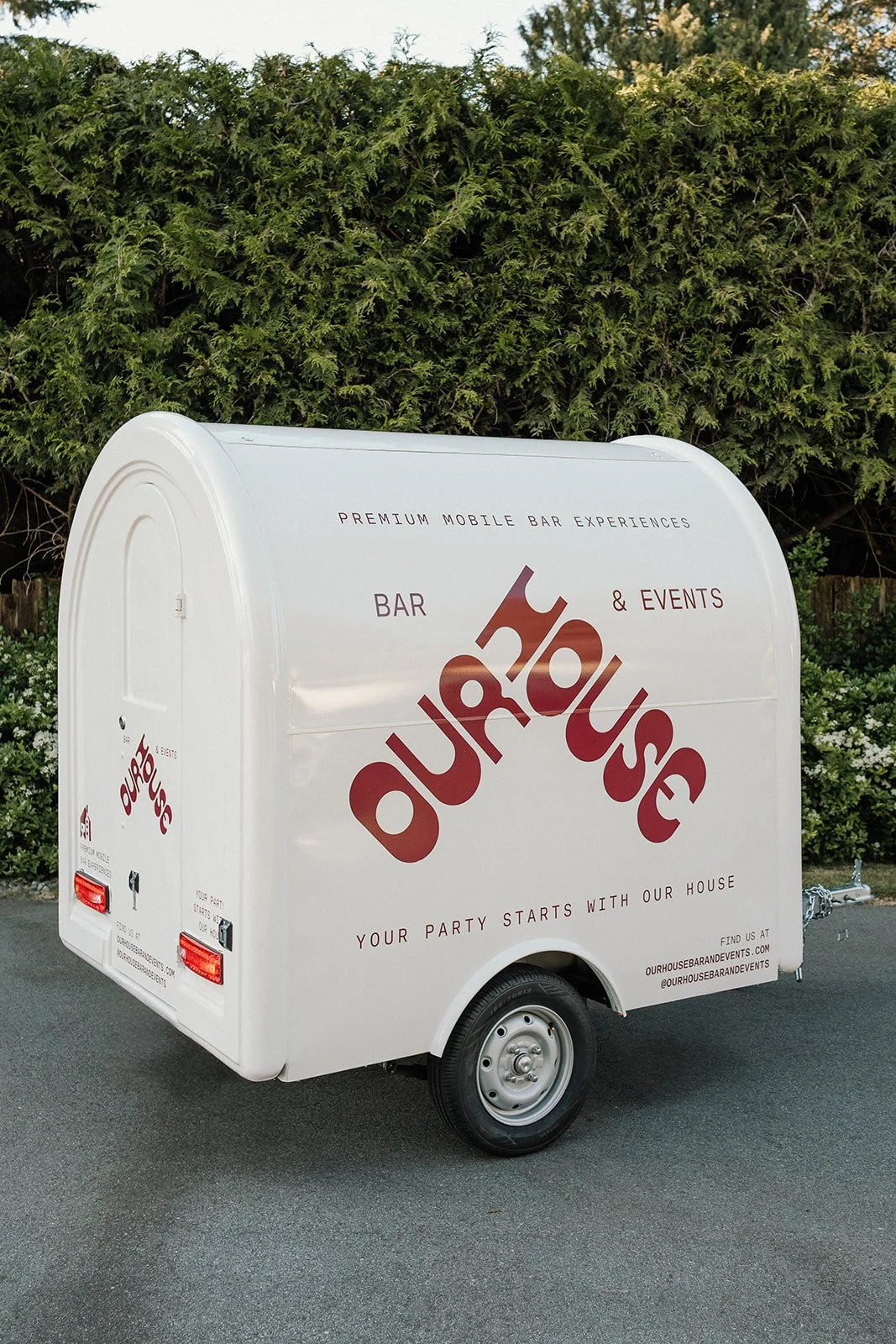 White mobile bar trailer with the logo 'ourhouse' in large red letters, parked on gray pavement with green shrubbery in the background.