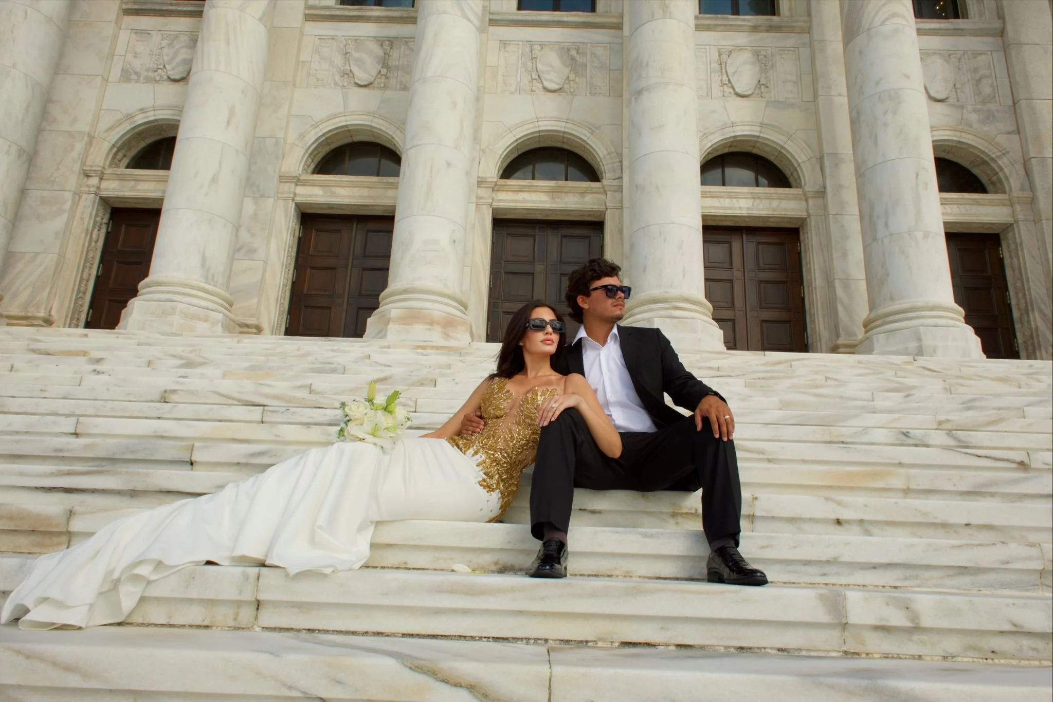 A couple dressed in formal attire sitting on marble steps in front of a grand classical building with tall columns and large wooden doors. The woman wears a white and gold dress with a bouquet of white flowers beside her, and the man wears a black su