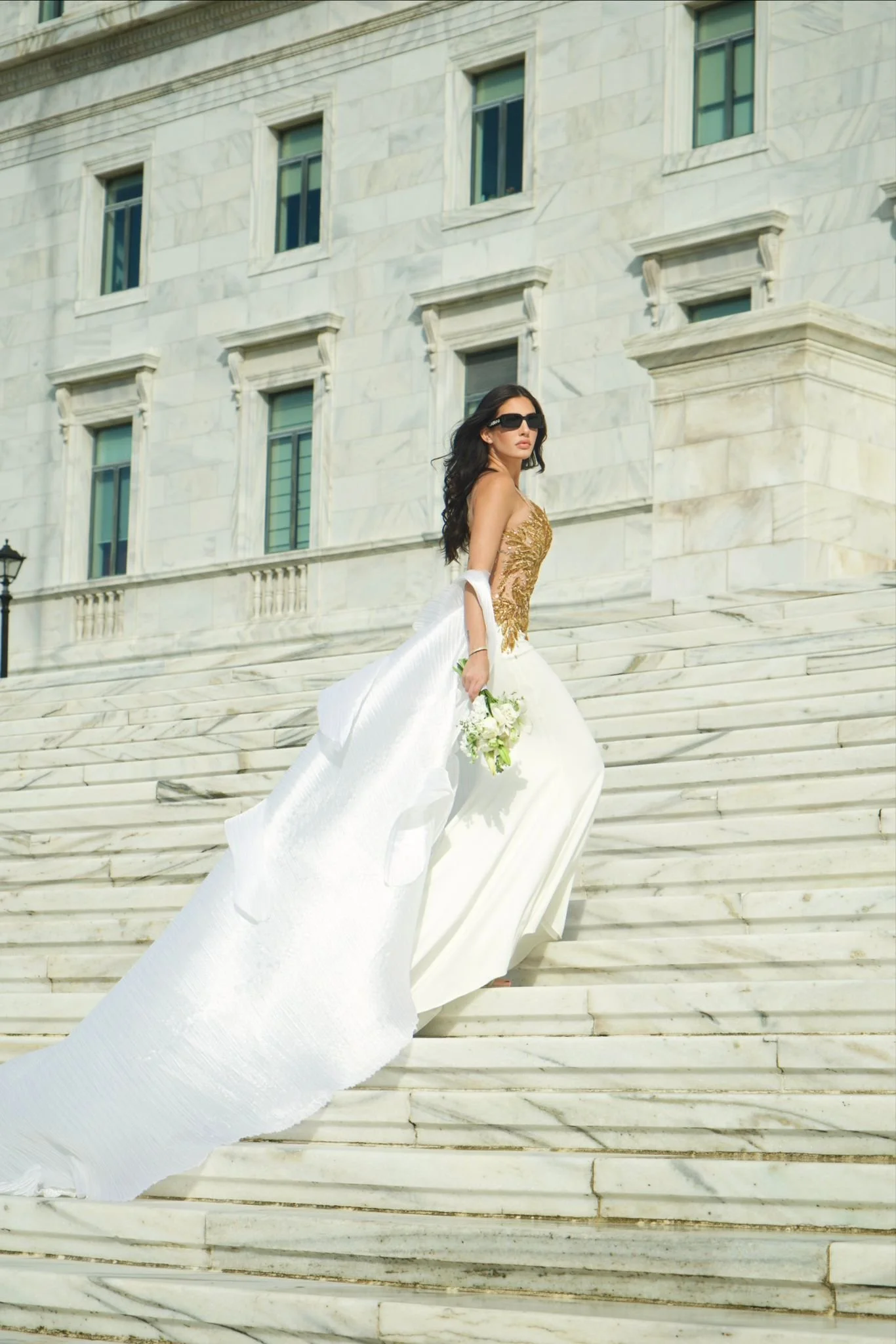 Woman in a wedding dress holding a bouquet, standing on marble stairs outside a large stone building.