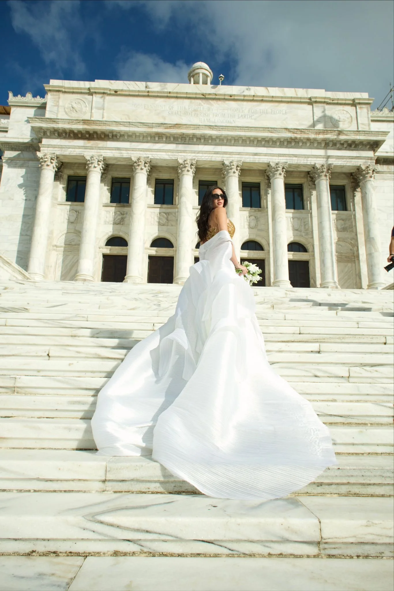 A woman in a white dress and sunglasses stands on the marble steps of the Lincoln Memorial in Washington, D.C.