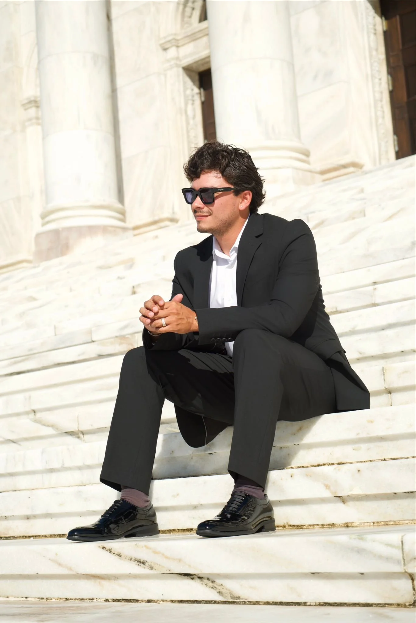 A man wearing a black suit, white shirt, sunglasses, and black shiny shoes sits on white marble steps in front of a historic building with large columns.