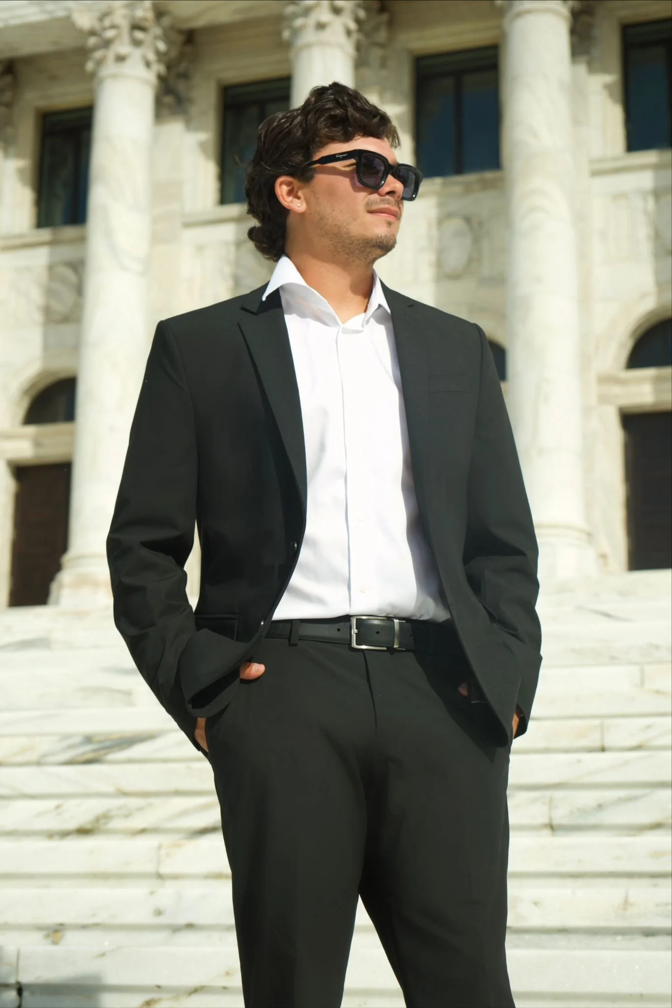 Man in black suit and white shirt wearing sunglasses stands outside a classical building with large columns.