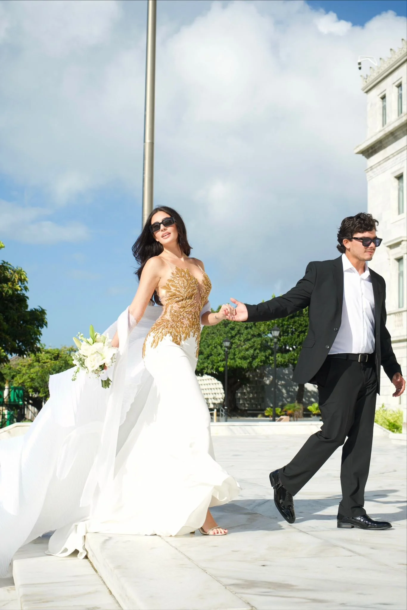 A woman in a wedding dress holding a bouquet is holding hands with a man in a tuxedo as they walk outdoors on a sunny day.
