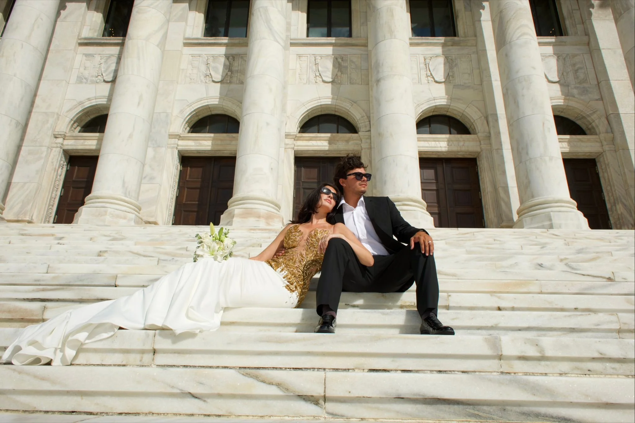 A newlywed couple sitting on the steps of a grand marble building, possibly a historic courthouse or government building, with large stone columns in the background. The bride is resting her head on the groom's shoulder, holding a bouquet of white fl