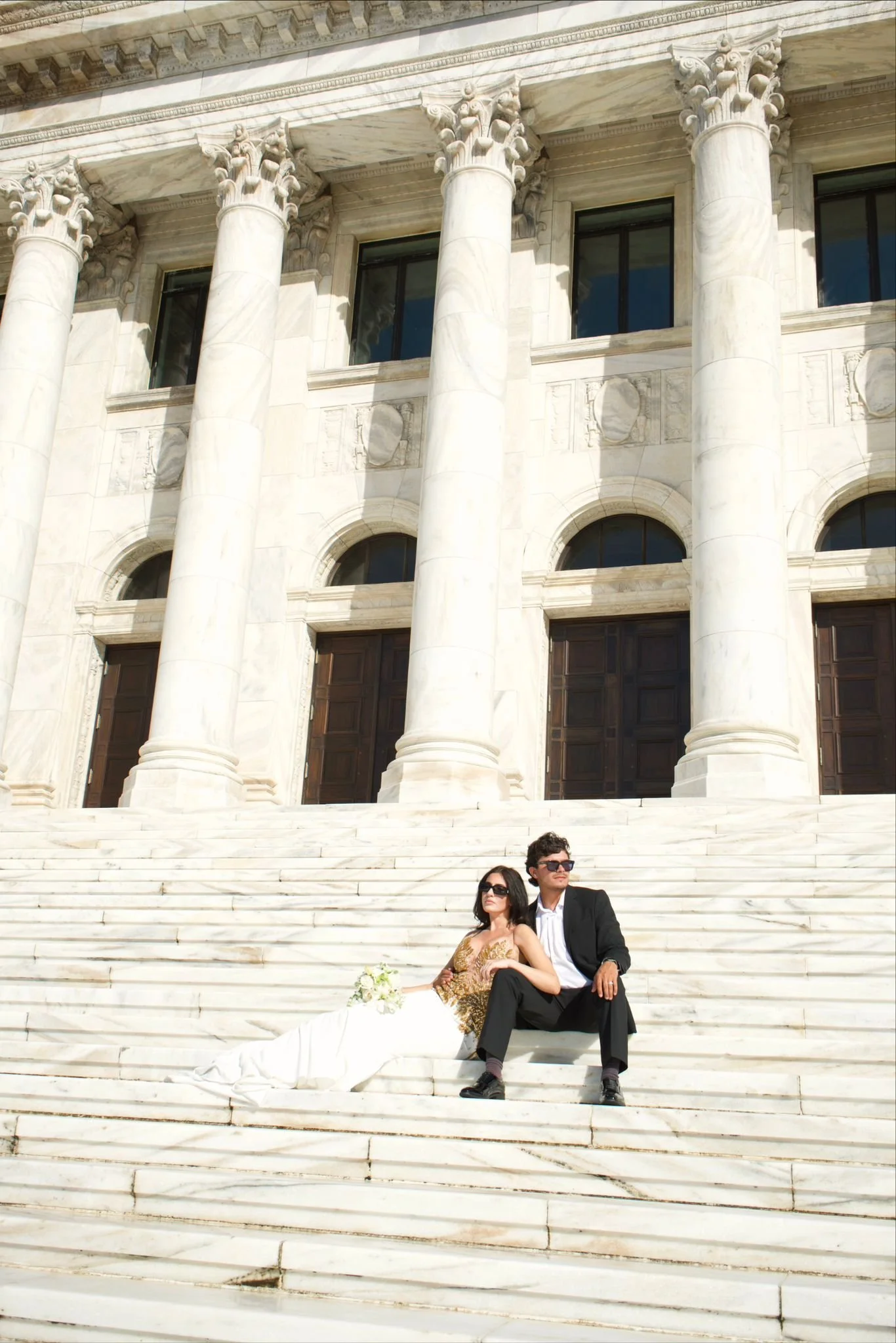 A couple in wedding attire sitting on white marble stairs in front of a grand building with large columns.
