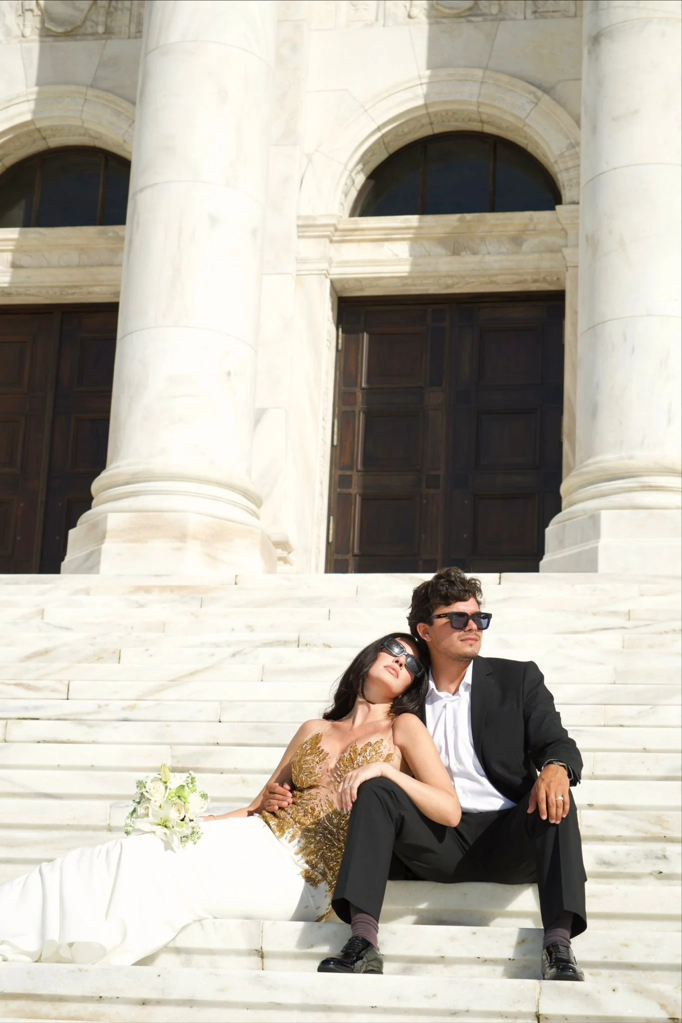 A stylish couple sitting and relaxing on white marble steps in front of a large classical building with tall columns and dark wooden doors. The woman is wearing a gold embellished dress and lying back with her head on the man's shoulder, holding a bo