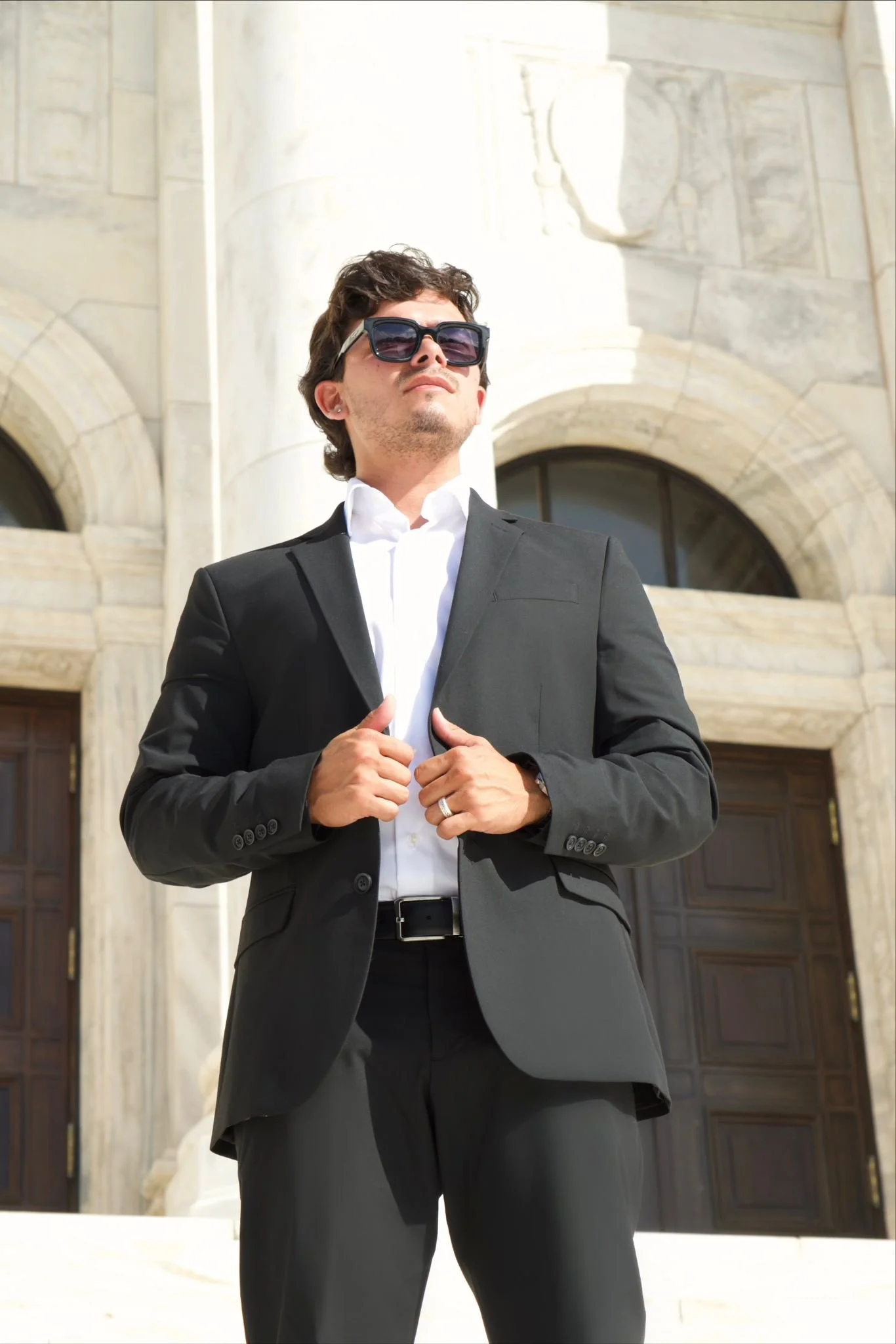 A man in a black suit and sunglasses standing in front of a historic building with large wooden doors.