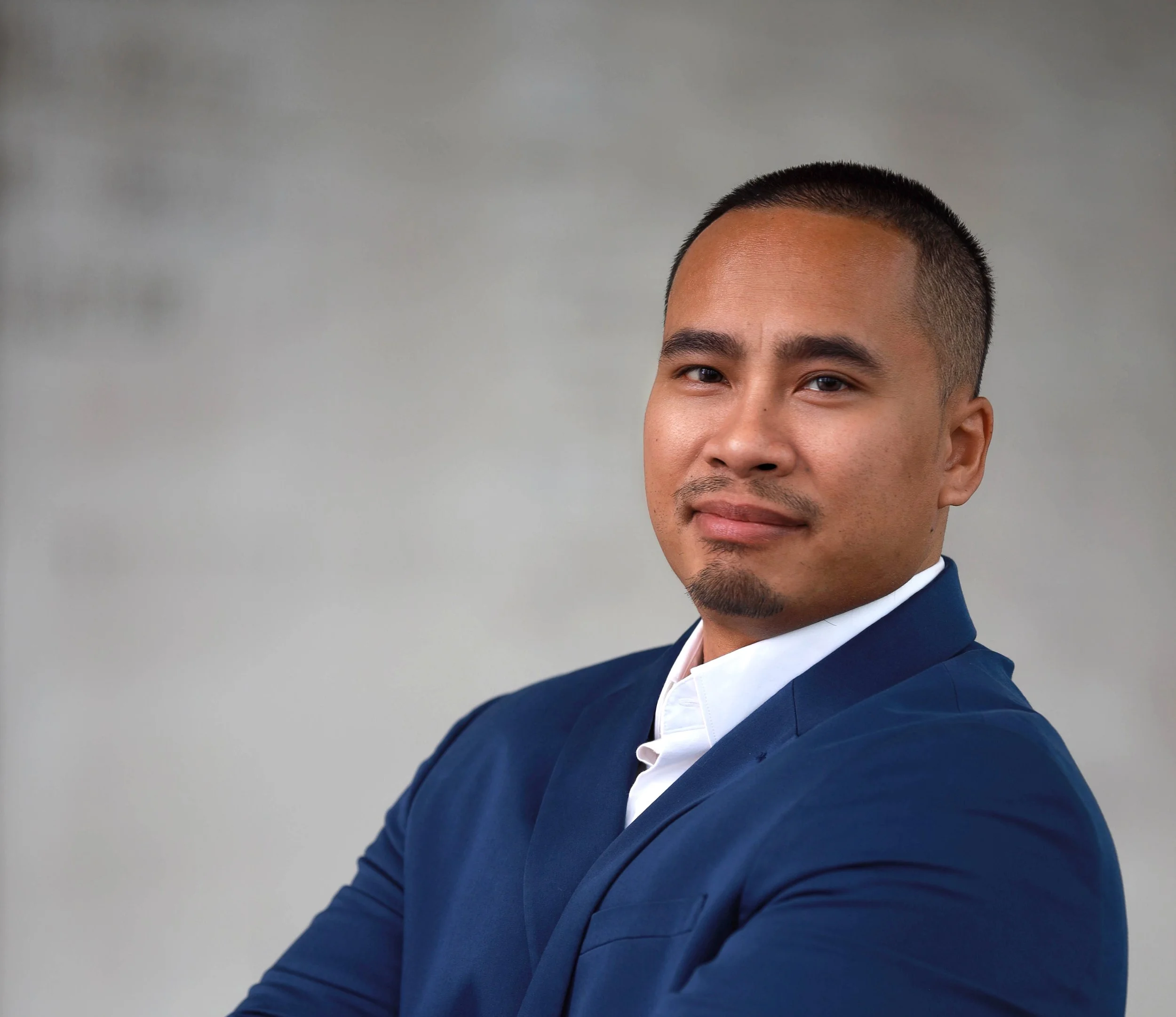 A man with short dark hair and a goatee wearing a navy blue blazer and white shirt, posing with crossed arms against a neutral background.