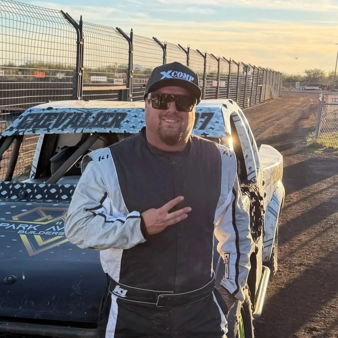A man in a racing suit and sunglasses making a peace sign, standing in front of a race car, at a dirt race track during sunset.