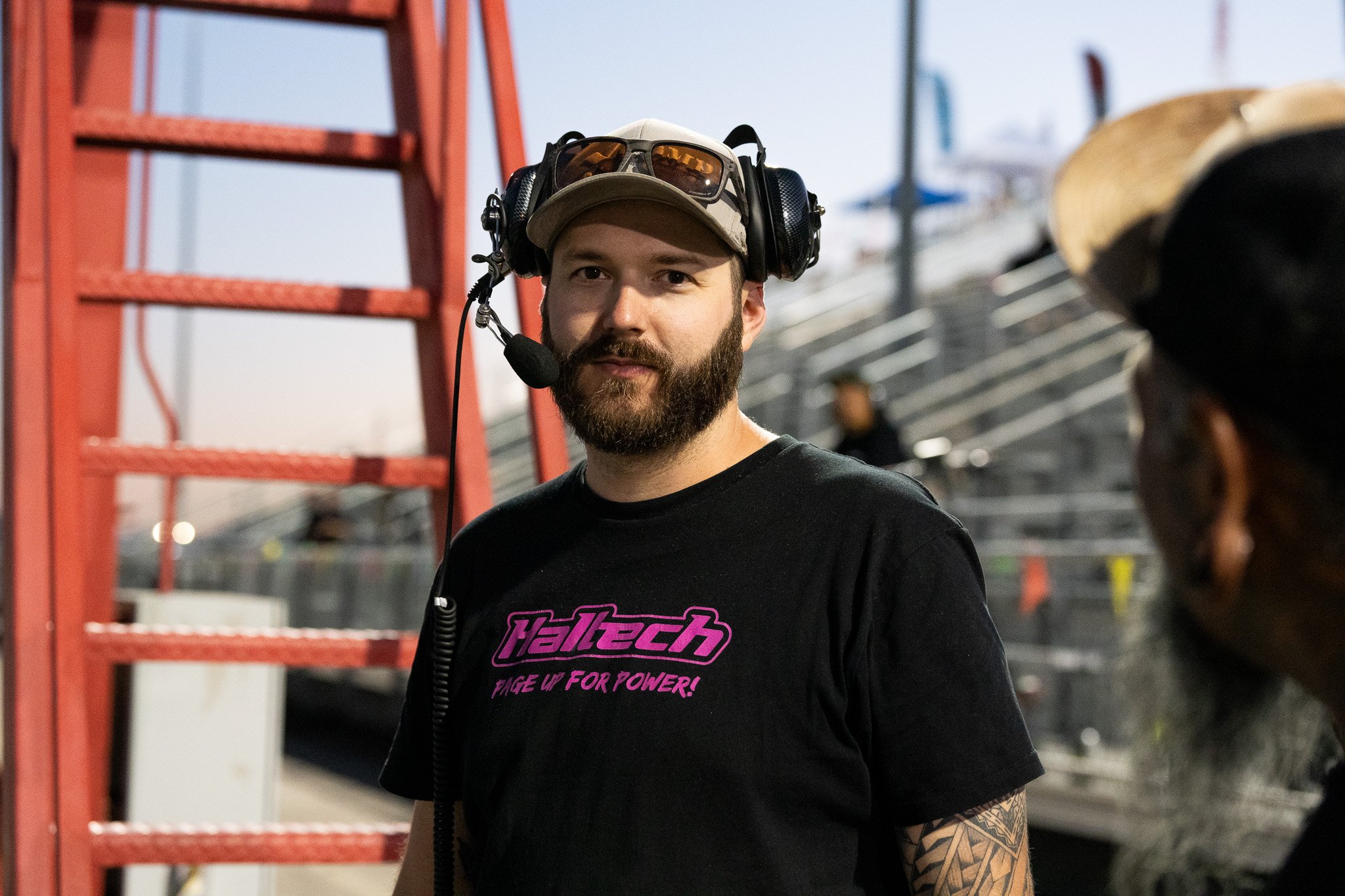 A man with a beard wearing a beige cap, black t-shirt with purple text, and large black headphones with a microphone, standing outdoors near a red ladder and bleachers, talking to another person.