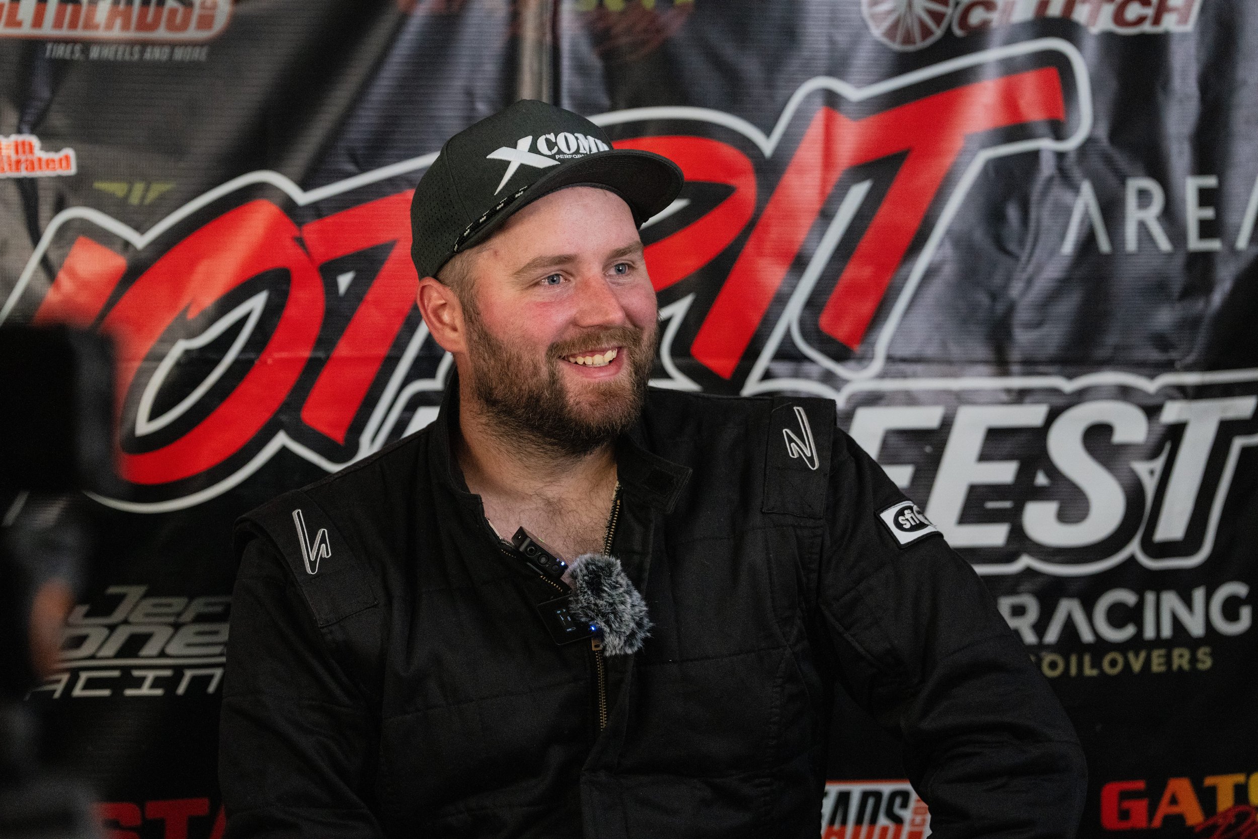 A smiling man with a beard and wearing a black cap and racing suit is sitting in front of a backdrop with NASCAR and racing branding.