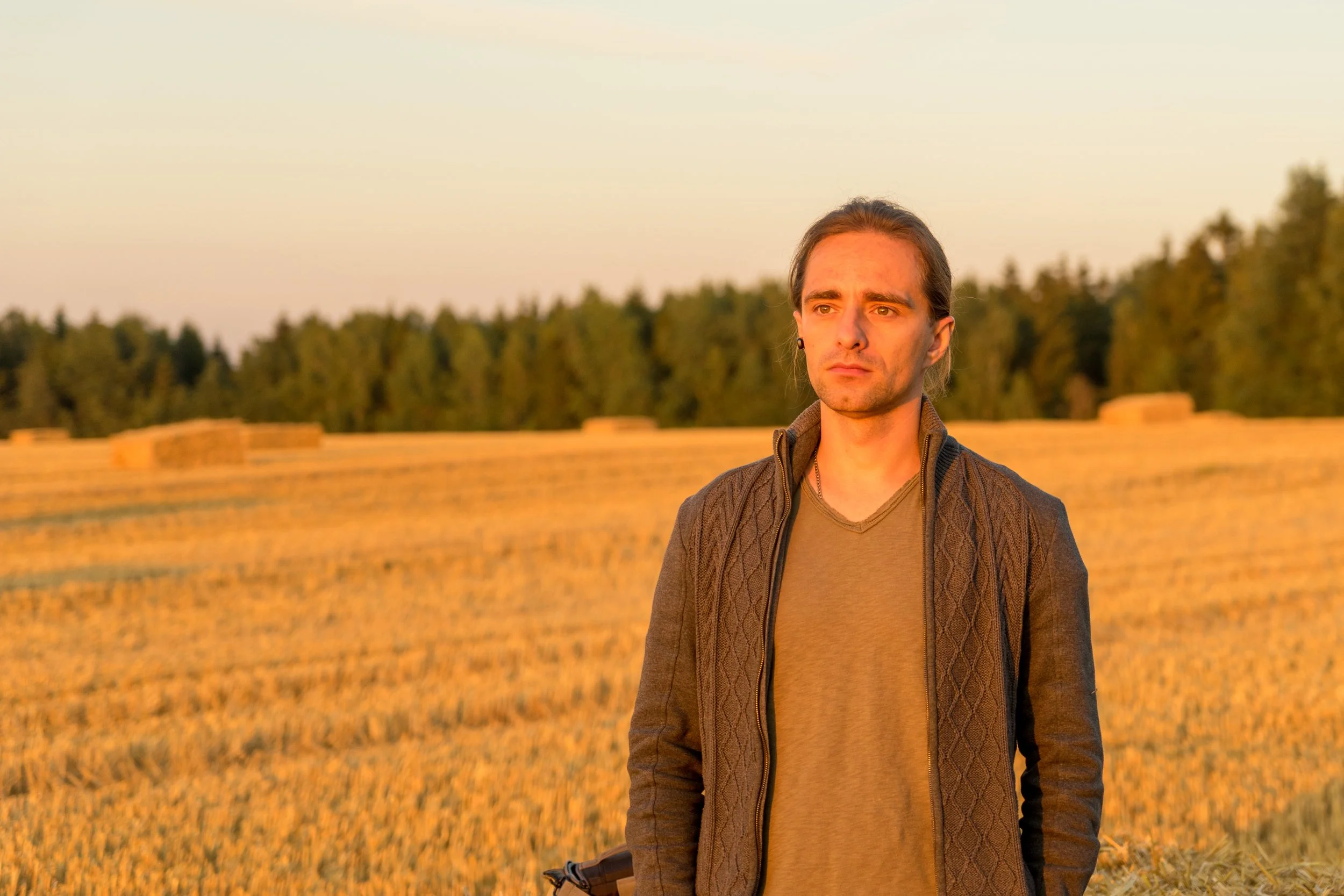 A man with long hair, wearing a brown jacket and T-shirt, standing outdoors in a field during sunset with hay bales and trees in the background.