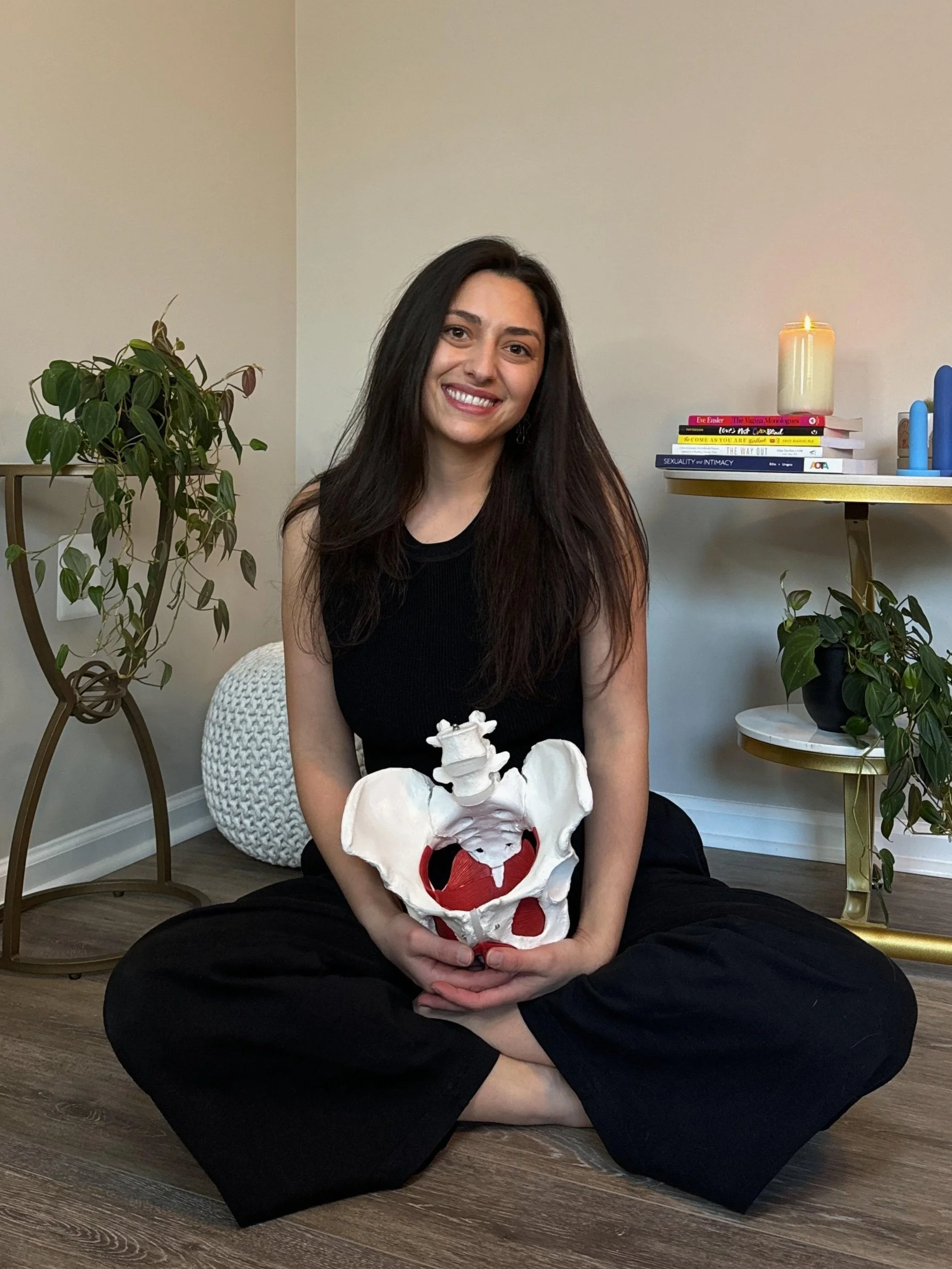 Gina, a pelvic floor therapist, in black holding pelvis/pelvic floor model smiling with dilators and pelvic & sexual health books behind her.