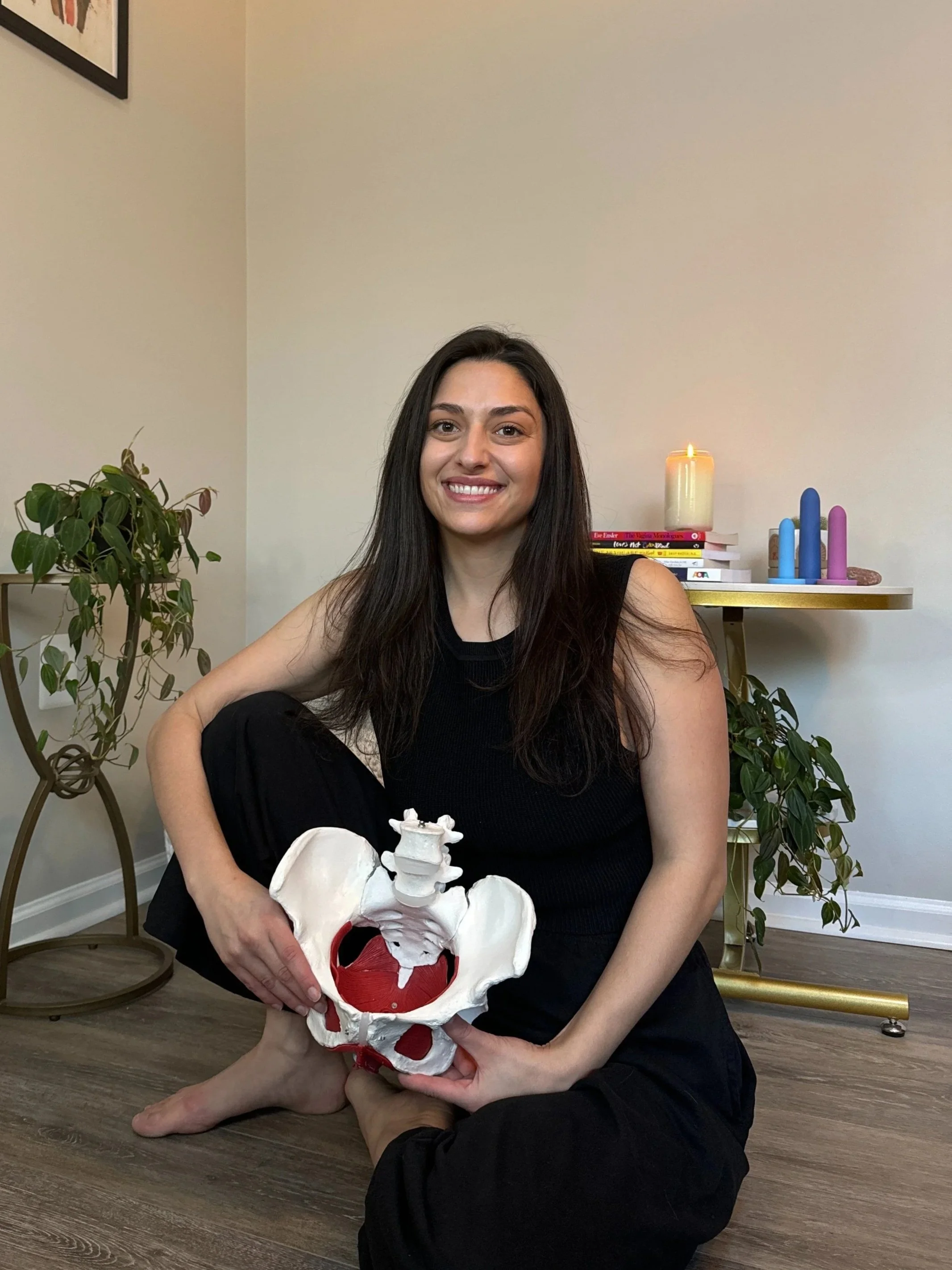 A young woman with long dark hair and a black sleeveless top sitting on the floor, holding a model of a human pelvis, in a room with houseplants and a side table with candles and books. gina inglese. pelvic floor therapist. denver colorado.