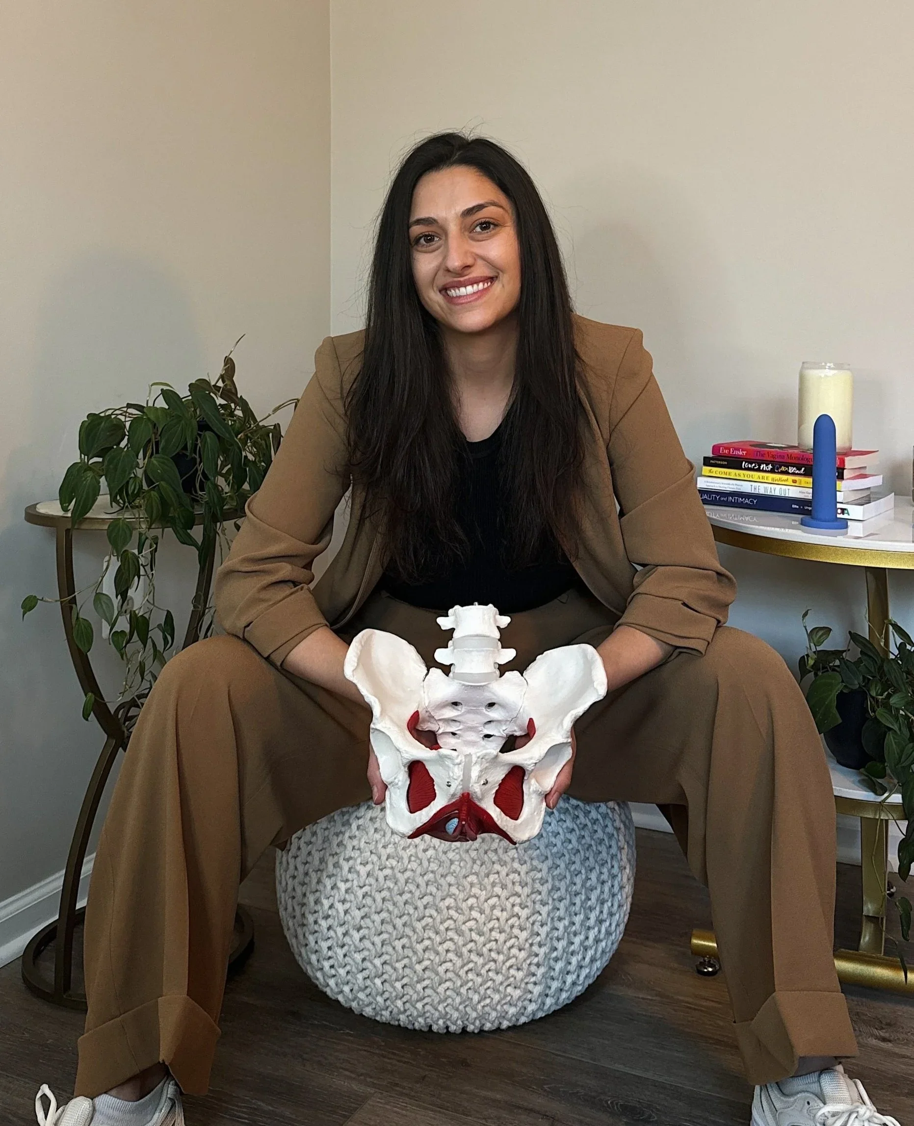A woman sitting on a knitted pouf, holding a model of a pelvis, in a room with plants, books, and decorative items on a side table.