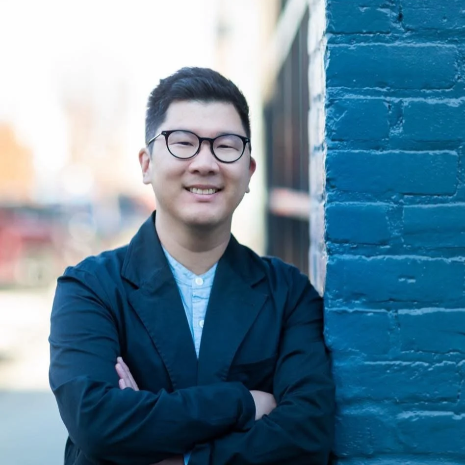 A smiling man with glasses standing next to a blue brick wall.