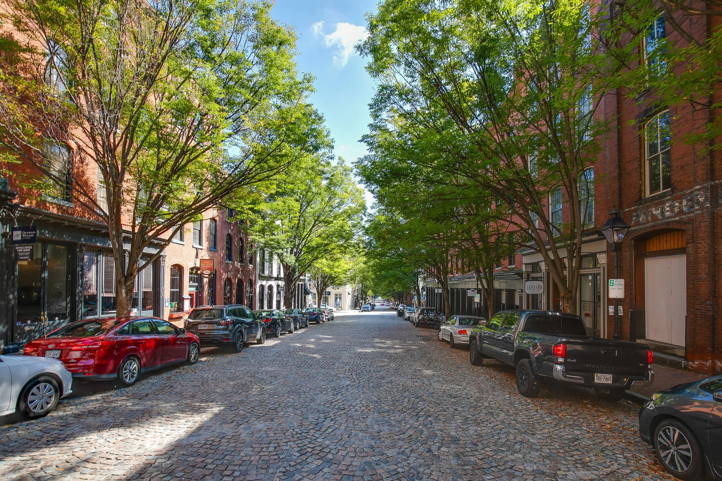 A cobblestone street lined with parked cars and trees on both sides, with multi-story brick buildings and storefronts, on a sunny day with a partly cloudy sky.