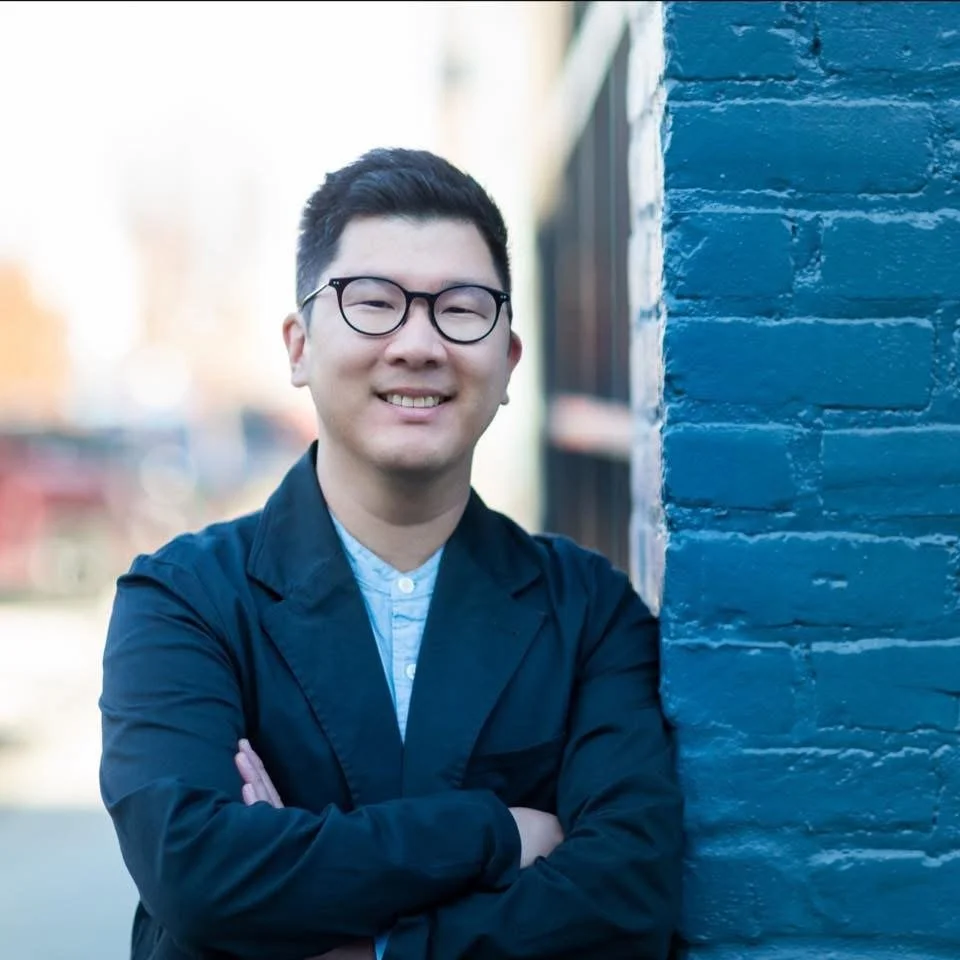 A smiling Asian man with glasses and black hair, wearing a navy blue jacket and a light blue shirt, standing outdoors next to a blue-painted brick wall.