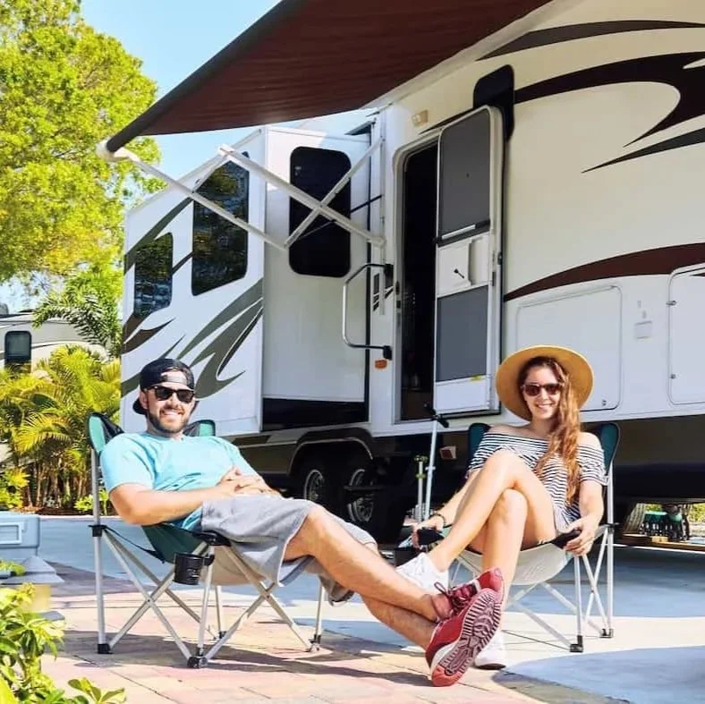 A man and woman relaxing outside a travel trailer, sitting in camping chairs, smiling, with a tree and blue sky in the background.