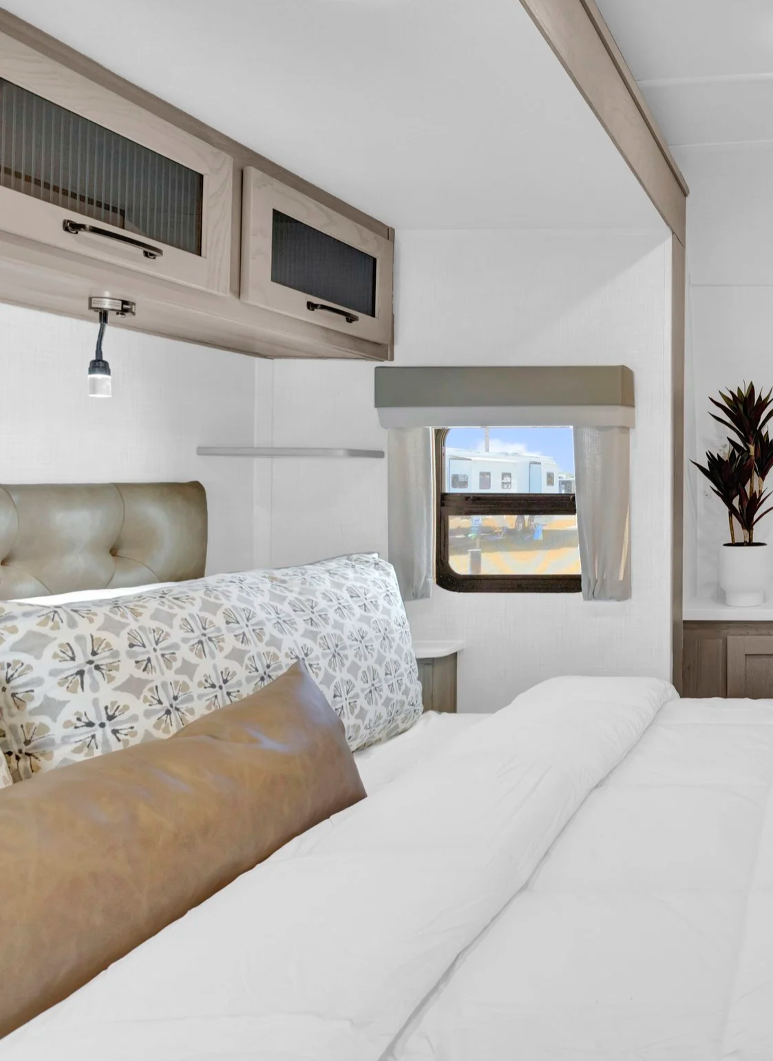 Interior of a bedroom with a window showing a trailer outside, a bed with patterned bedding and brown accent pillows, a potted plant, and overhead cabinets.
