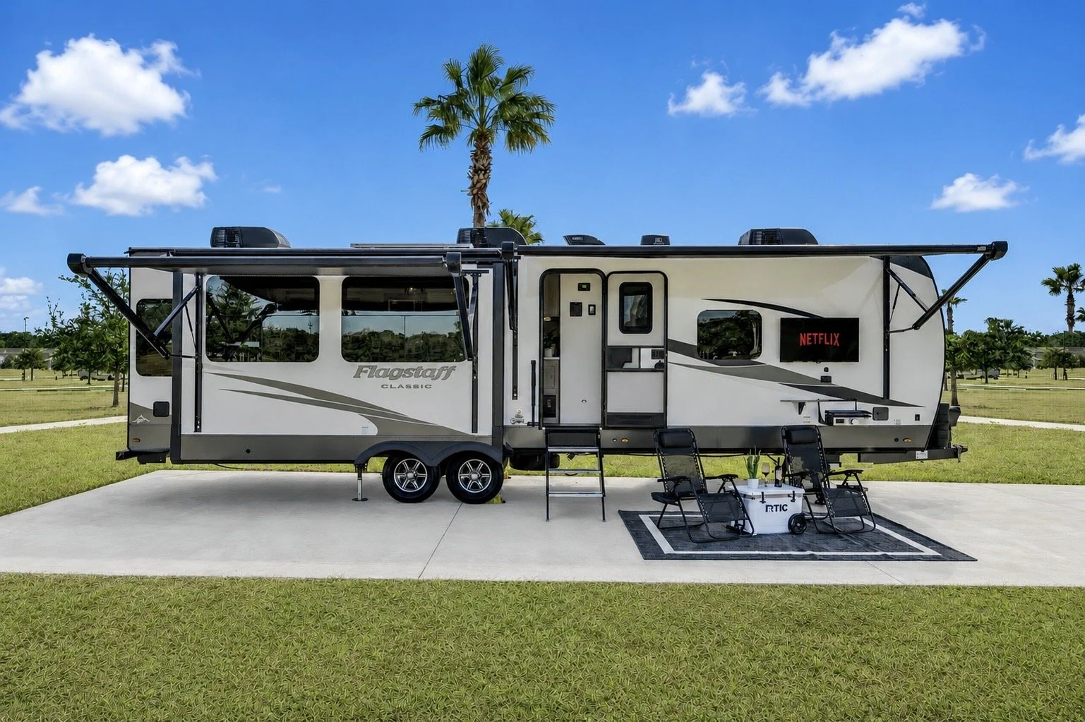 Living room inside a recreational vehicle with a brown sofa, window view of an outdoor field, mounted TV showing a beach scene, and decorative elements.