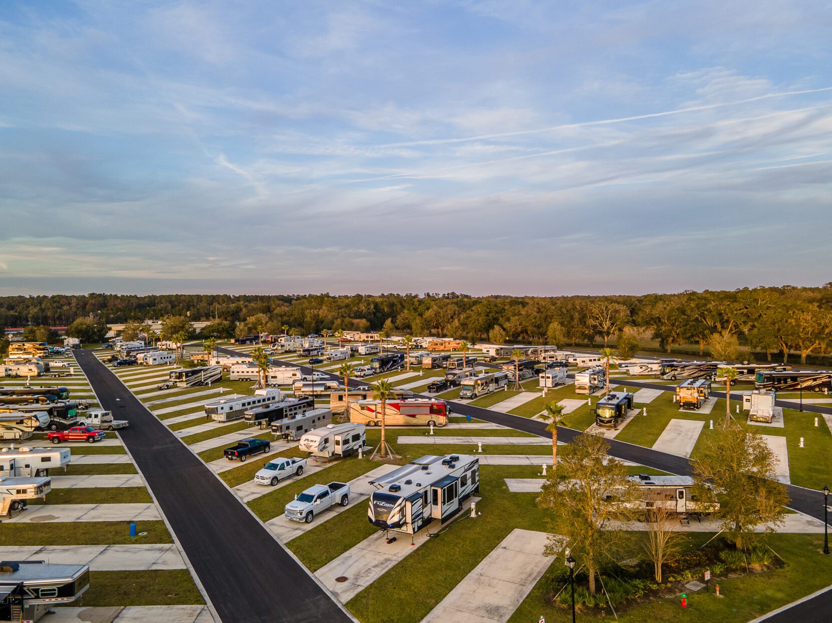 An aerial view of a large RV and camper parking lot with neatly arranged spaces, surrounded by a few trees and vehicles, under a partly cloudy sky during sunset.