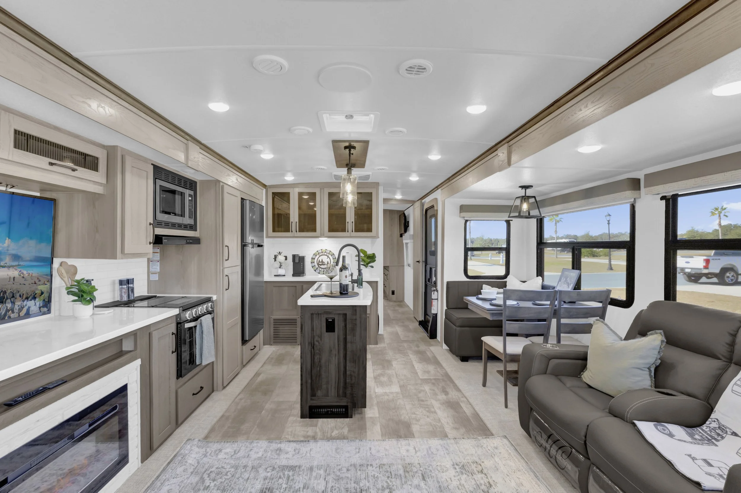 Interior of a modern, well-lit camper with a kitchen and living area, featuring a dining table, sofa, and large windows showing an outdoor street scene.