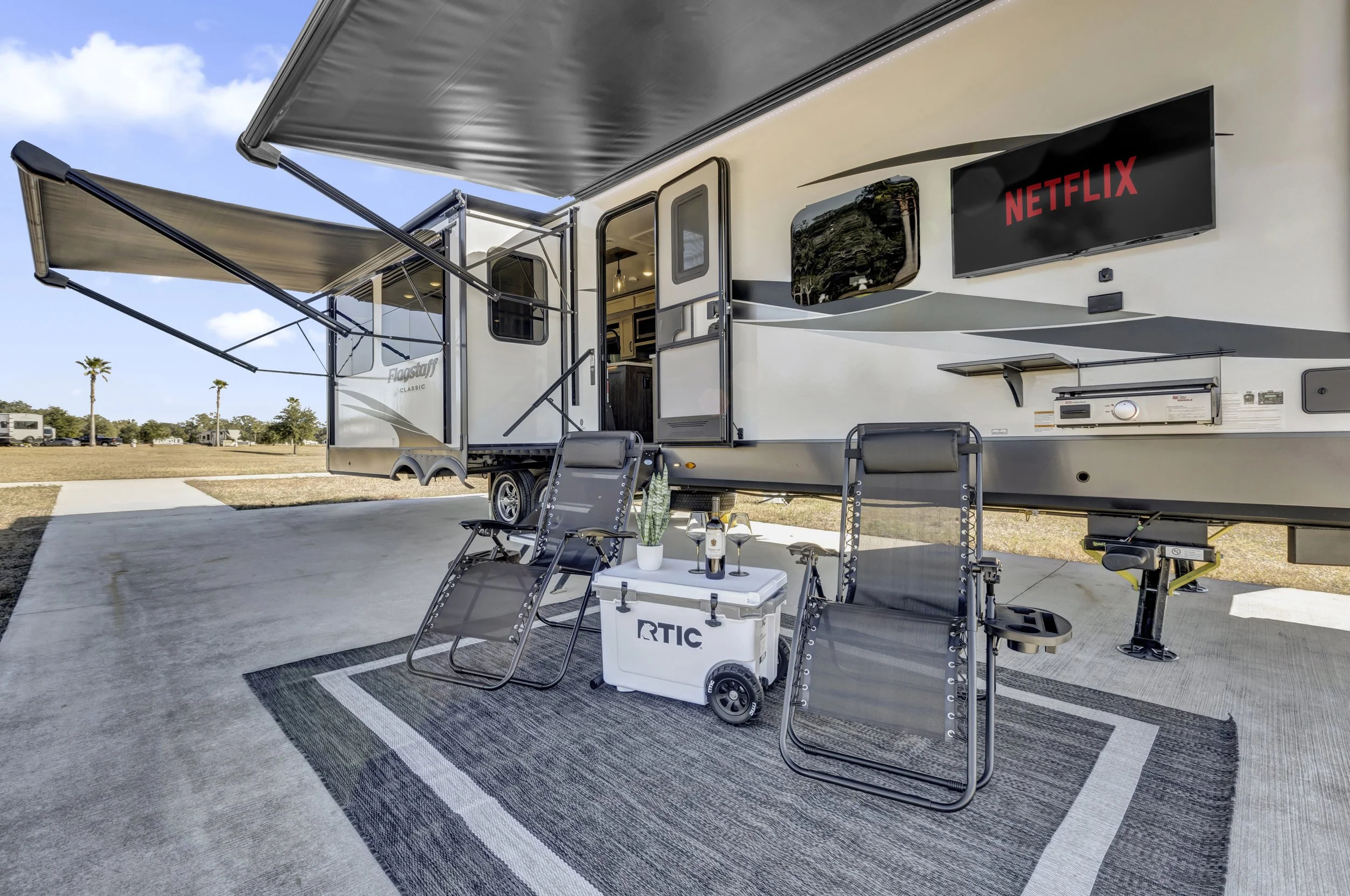Outdoor scene featuring a modern RV with an extended awning, two lounge chairs, a small table with drinks, a potted plant, and a portable cooler, set on a concrete pad with a sunset or sunrise sky in the background.