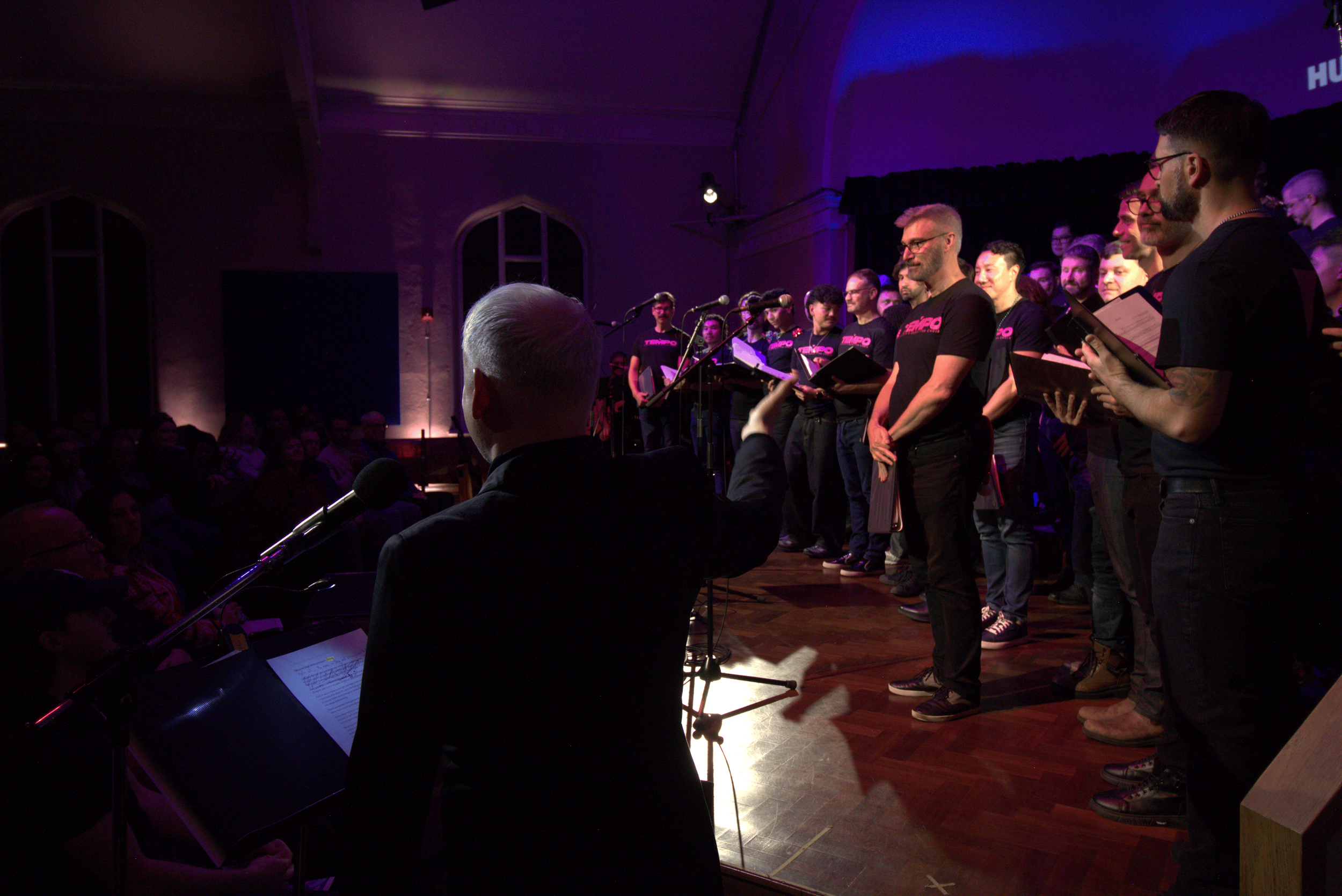A choir or singing group performing on stage in front of an audience, with a conductor or director leading them, in a dimly lit venue.