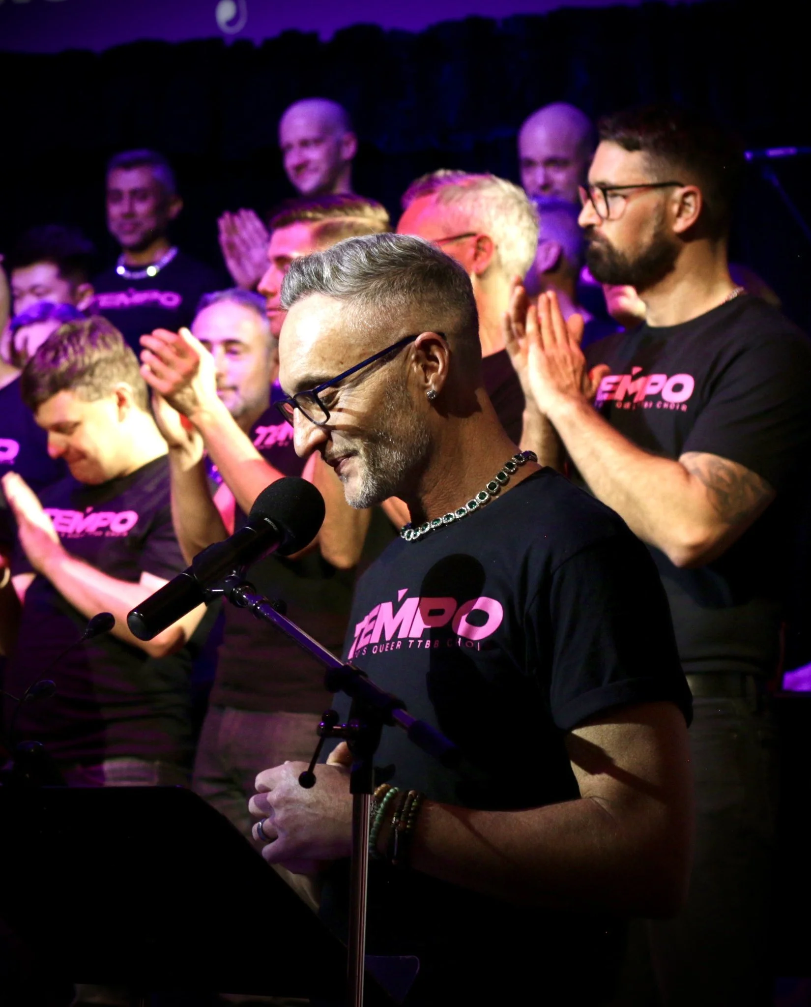 Group of people on stage wearing black shirts with pink text, some with their hands clasped, one man speaking into a microphone with glasses and jewelry, under stage lighting.