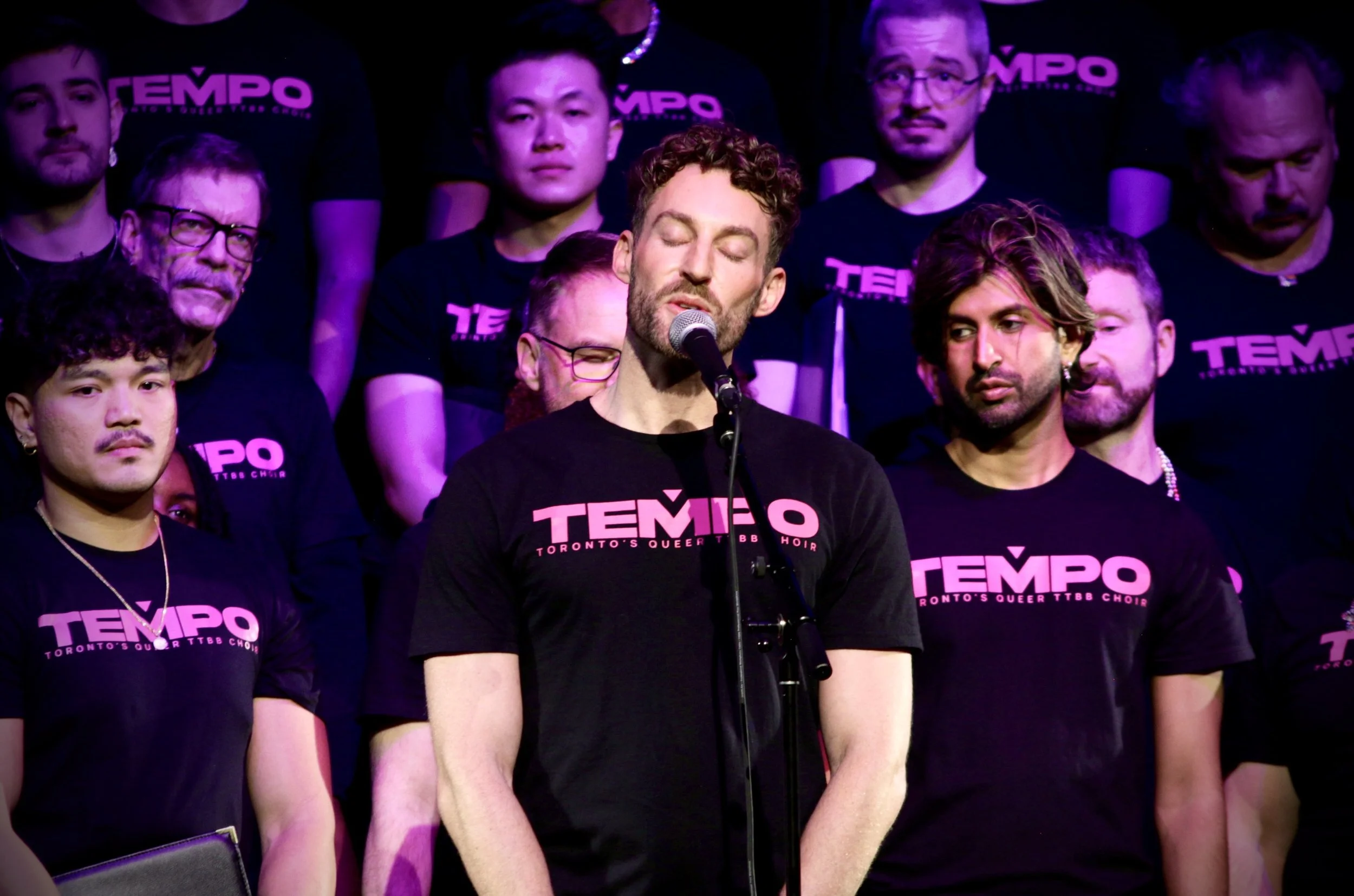 A group of people on stage, wearing black T-shirts with pink text that reads 'TEMPO Toronto's Queer TTBB Choir.' One person at the front has eyes closed and is singing into a microphone.