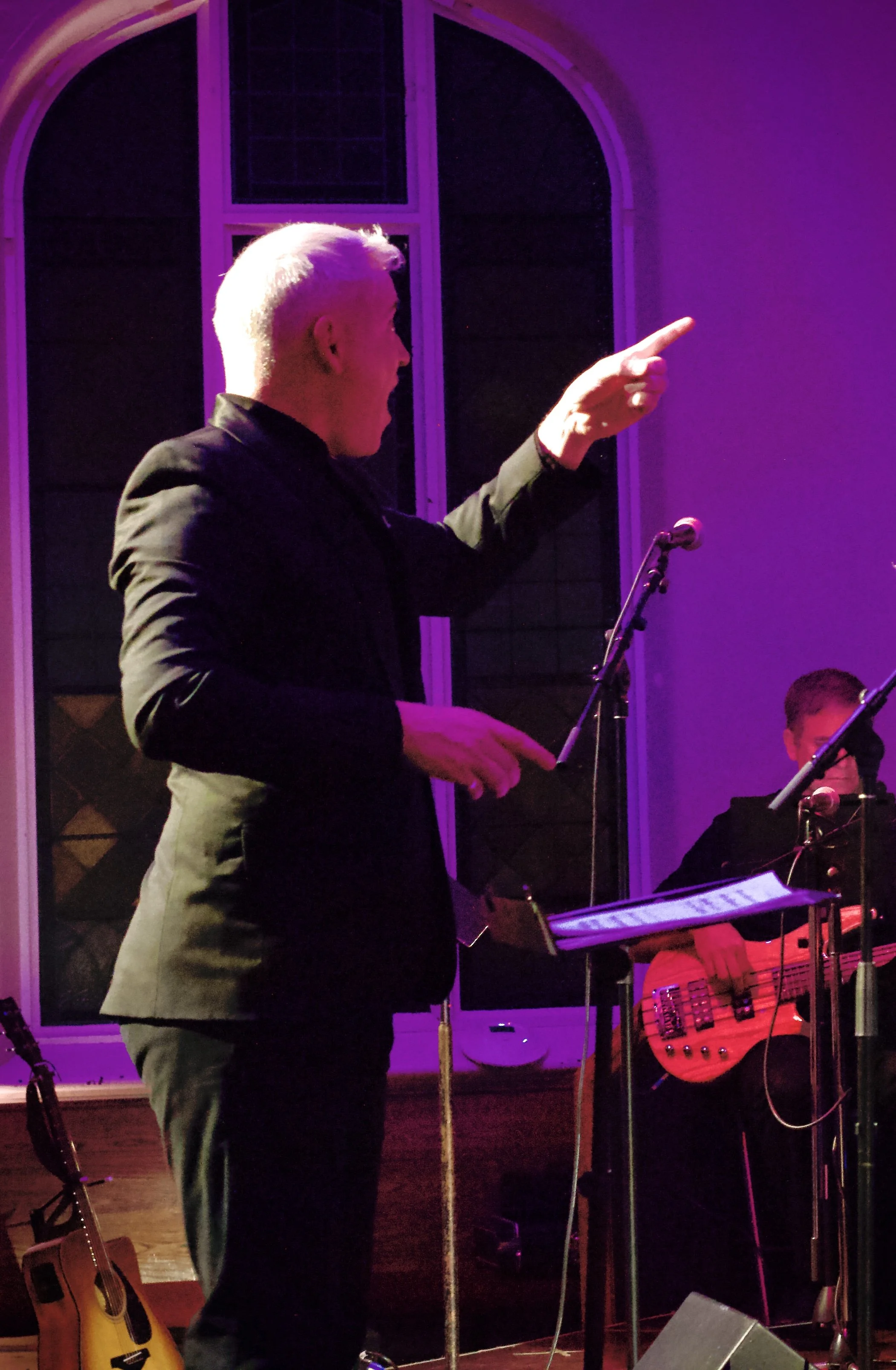 A man with gray hair wearing a black suit is standing on stage, pointing with his right hand, while holding a sheet of paper in his left hand. Behind him, a woman is sitting, playing a bass guitar, and there is a microphone stand in front of her. The stage is lit with purple lighting, with a large window in the background.