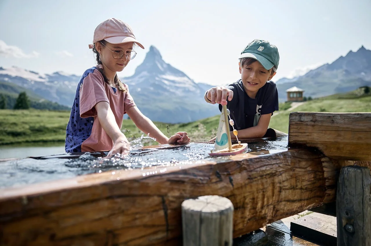 Two children, a girl and a boy, playing with a toy boat in a water trough outdoors with mountain scenery in the background.