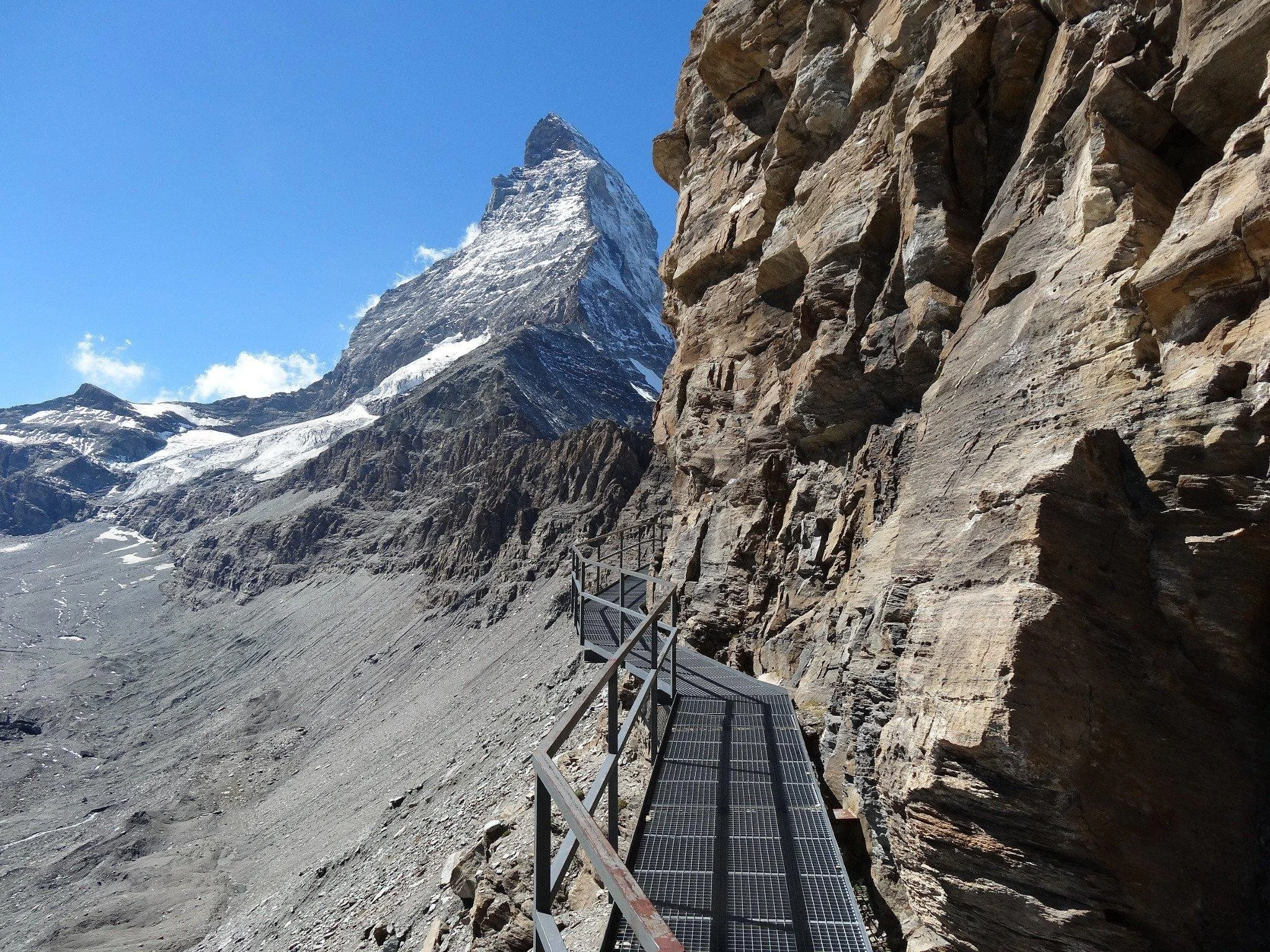 A narrow metal walkway with safety railings along a steep mountainside, with a snow-capped mountain peak in the background under a clear blue sky.