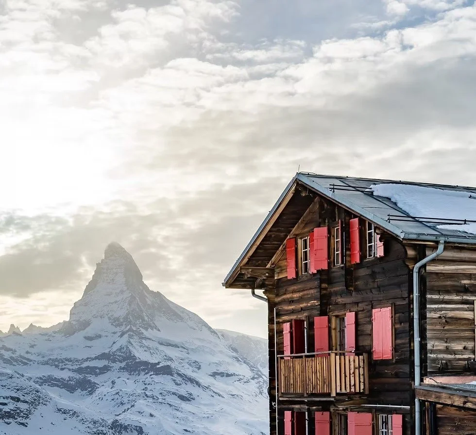 A wooden cabin with pink shutters on the windows, snow on the roof, and the Matterhorn mountain in the background, under a cloudy sky.