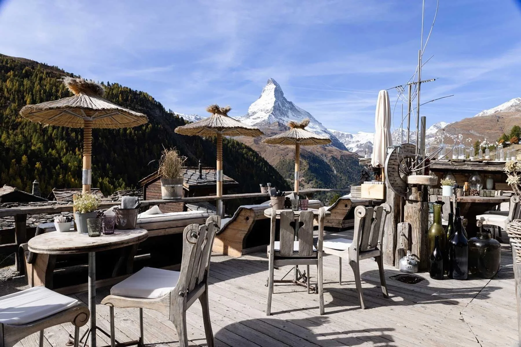 Outdoor terrace with wooden furniture and straw umbrellas, overlooking mountains with a snow-capped peak in the background.