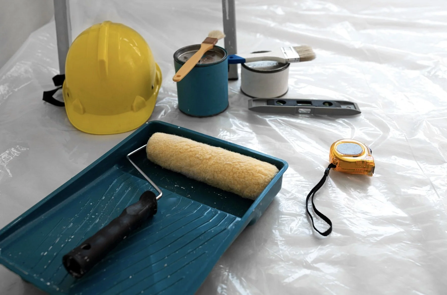 Construction tools including yellow hard hat, paint roller, paint cans with brushes, spirit level, measuring tape, and protective plastic sheeting on a surface.