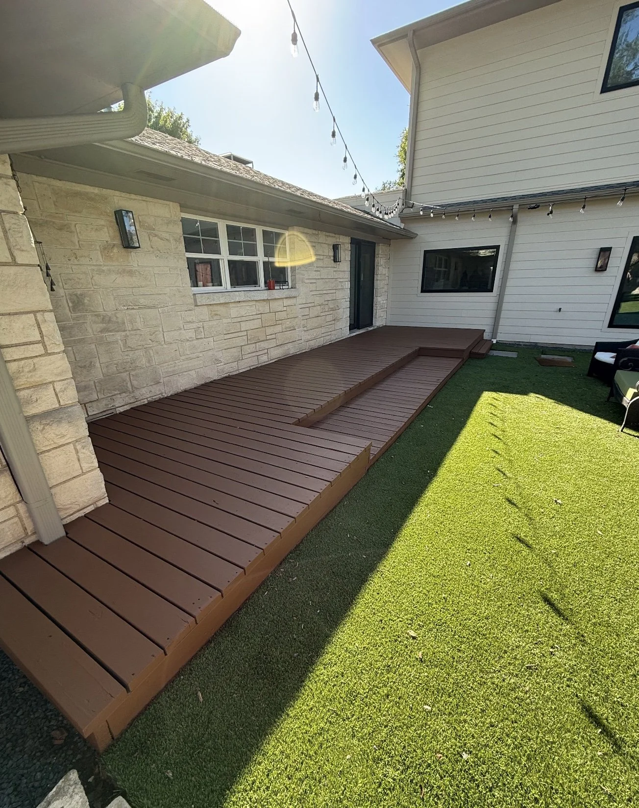 Backyard with wooden deck, green grass, and house walls with windows and siding, under a sunny sky with string lights overhead.