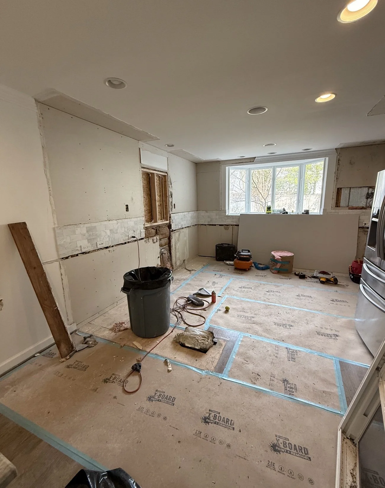 Kitchen under renovation with exposed drywall, construction tools, and flooring in progress, illuminated by recessed ceiling lights and a large window.