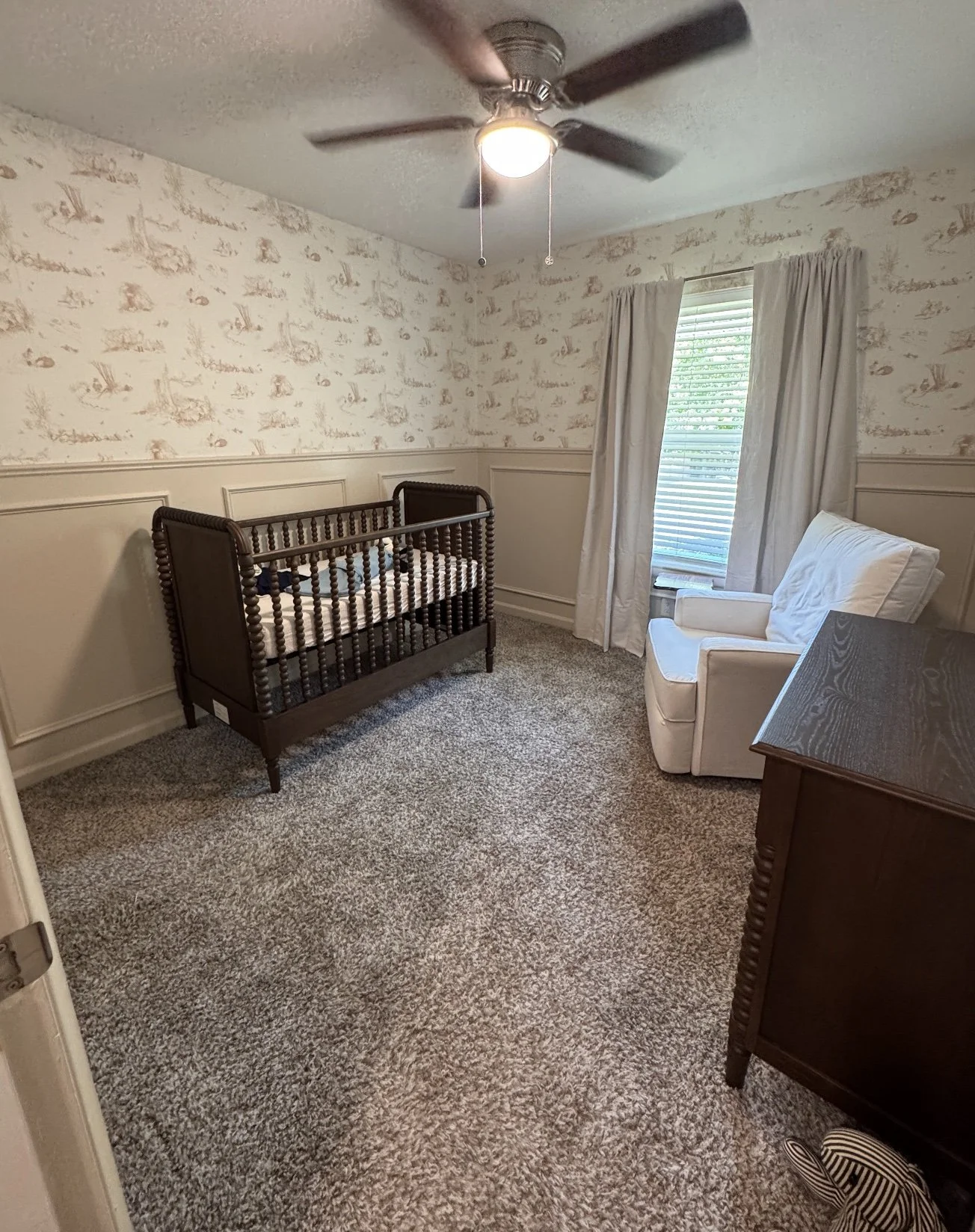 A nursery room with beige floral wallpaper, carpeted floor, ceiling fan with light, a brown wooden crib, white armchair, and a dark wooden dresser.