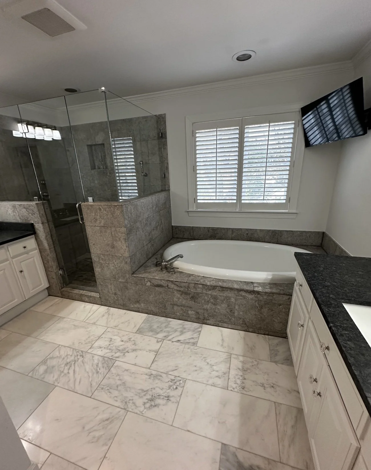 Bathroom with a glass-enclosed shower, bathtub, white cabinets, black countertop, and a wall-mounted TV.