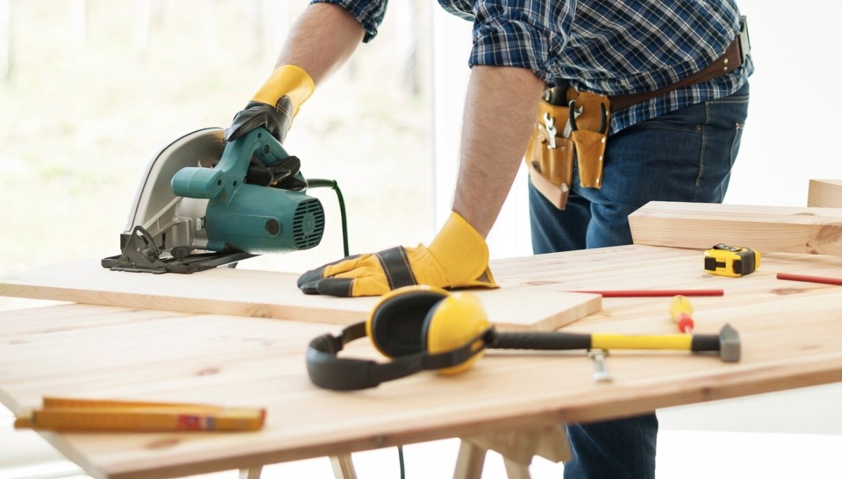 Person wearing yellow work gloves using a circular saw on a wooden board at a workbench.