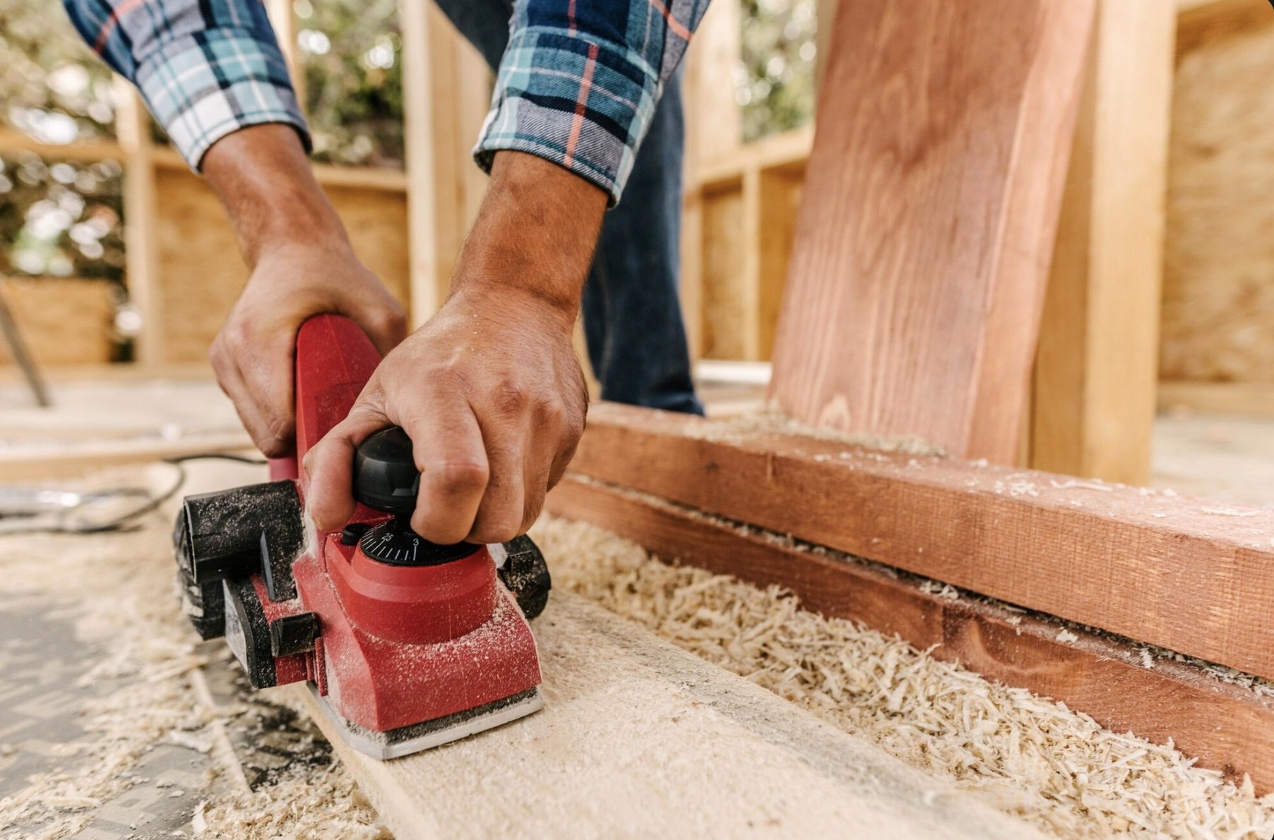 A person using a red electric sander to smooth a wooden plank at a construction site with wooden framing in the background.