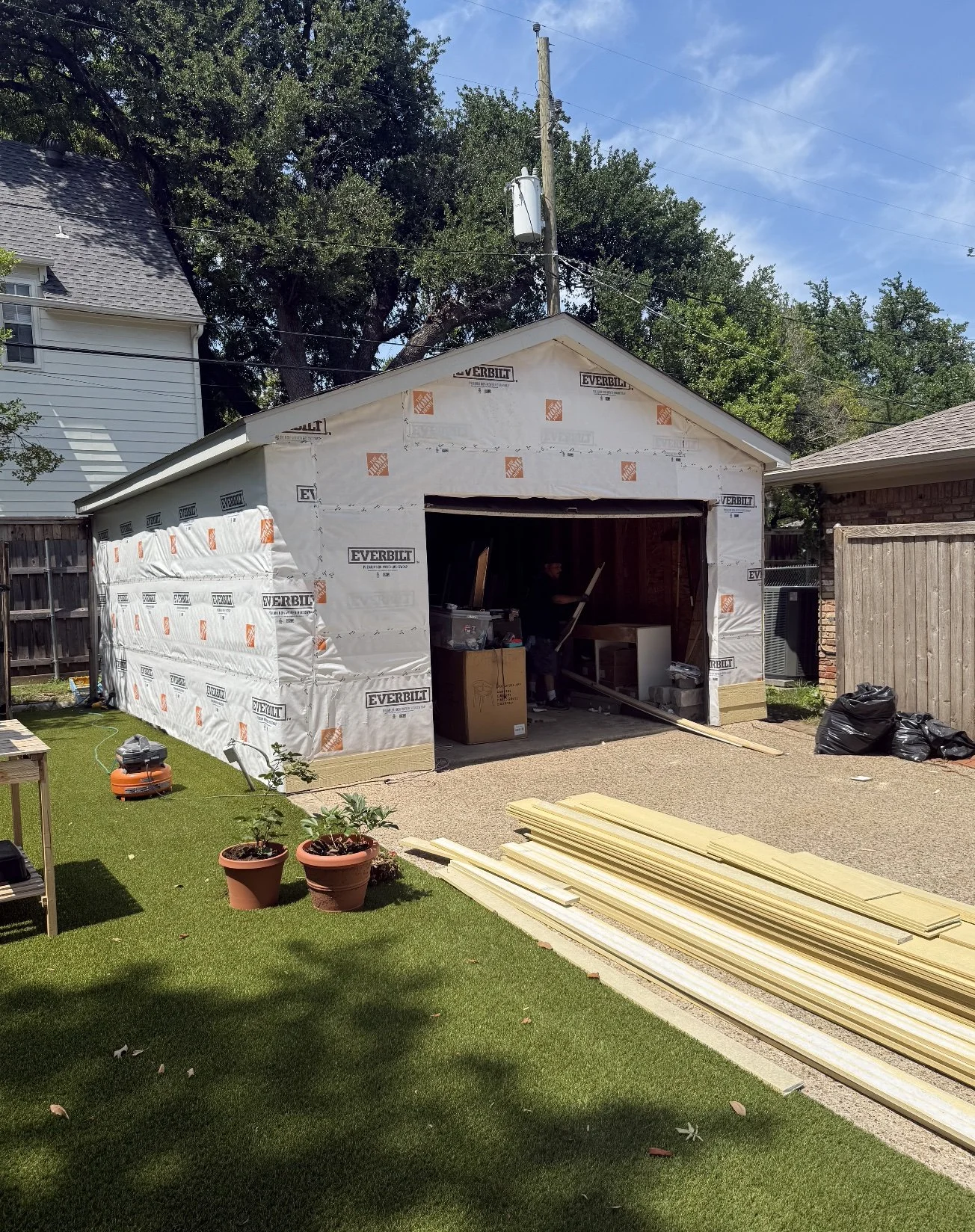 A garage under construction with white sheathing and building materials outside, including yellow wooden planks on the driveway, two potted plants, and a black trash bag, on a sunny day.