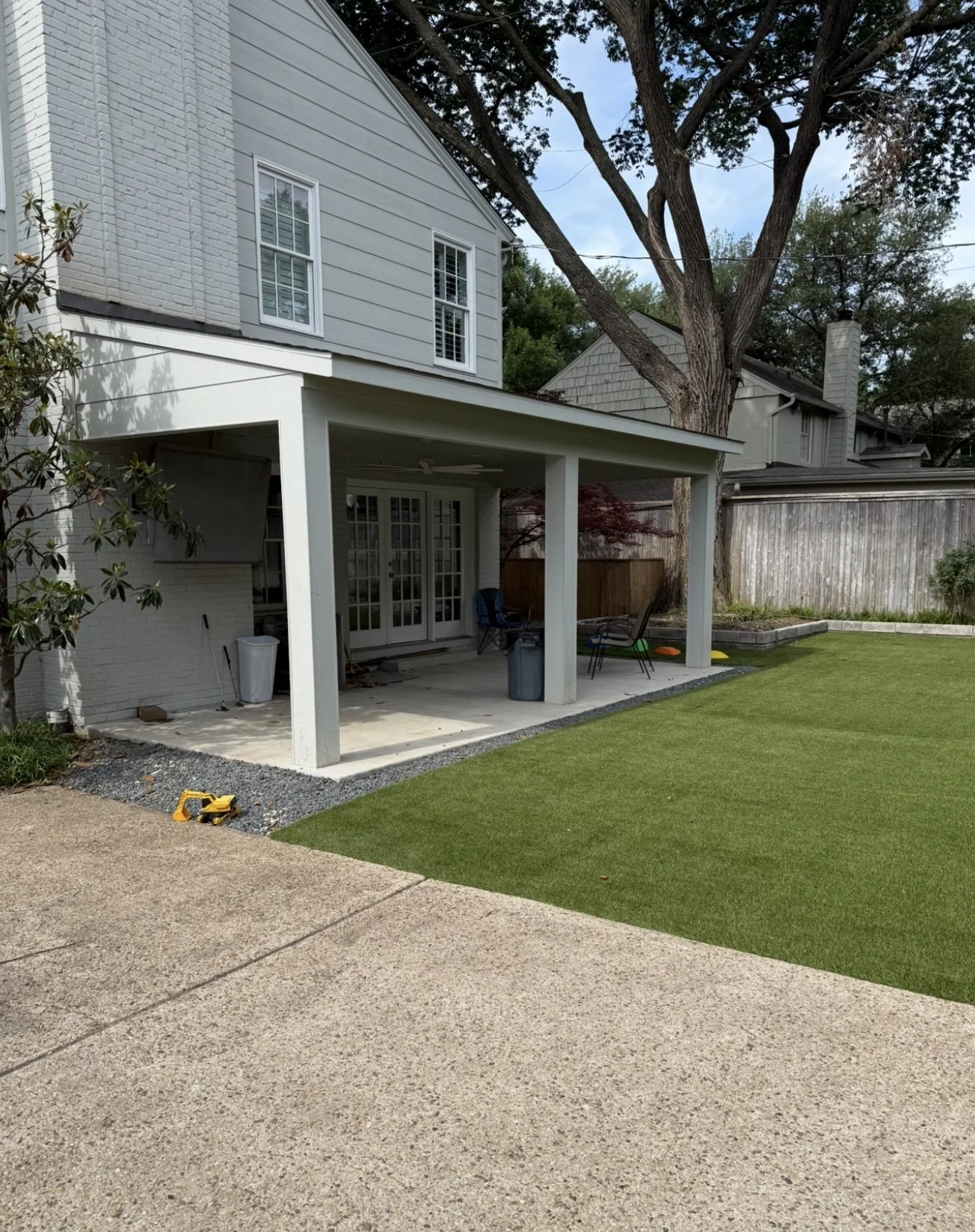 Backyard patio area with a covered porch, outdoor chairs, and a grassy lawn under a large tree, with fencing and neighboring houses in the background.