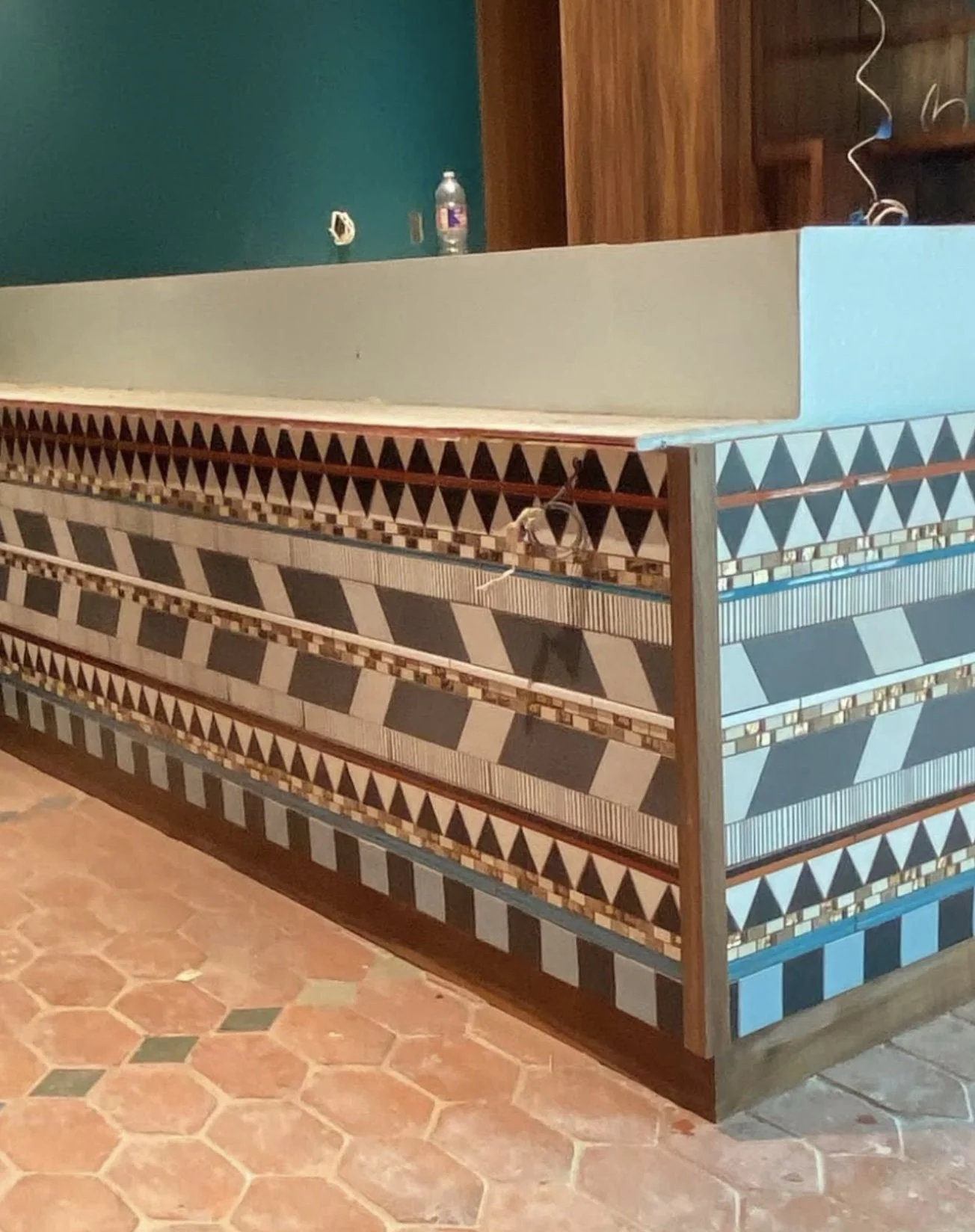 Close-up of a decorative wooden counter with geometric patterns in black, white, brown, and light blue, in a room with terracotta tile flooring and a teal-colored wall in the background.
