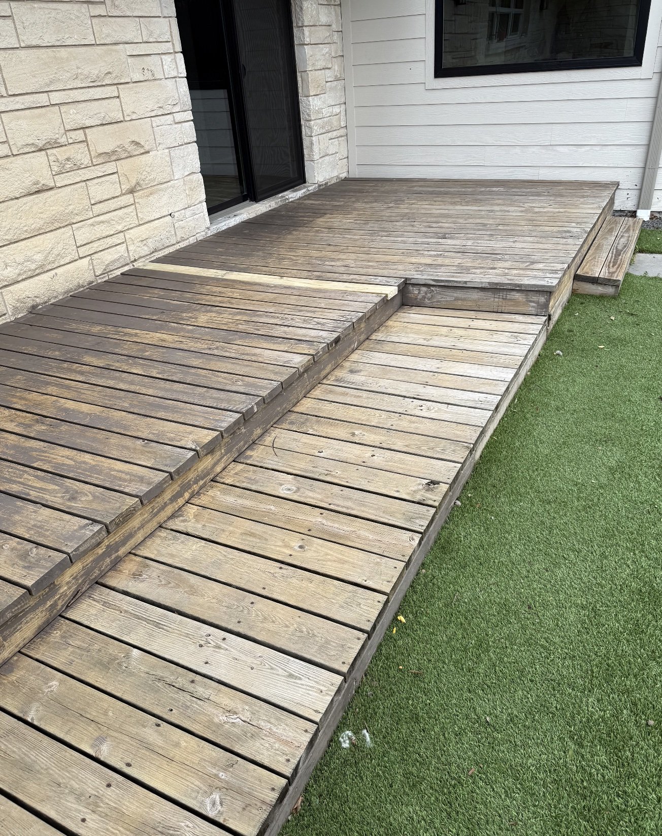 Wooden deck attached to a house with a sliding glass door. The deck has multiple levels with wooden planks and some grass visible beside it.