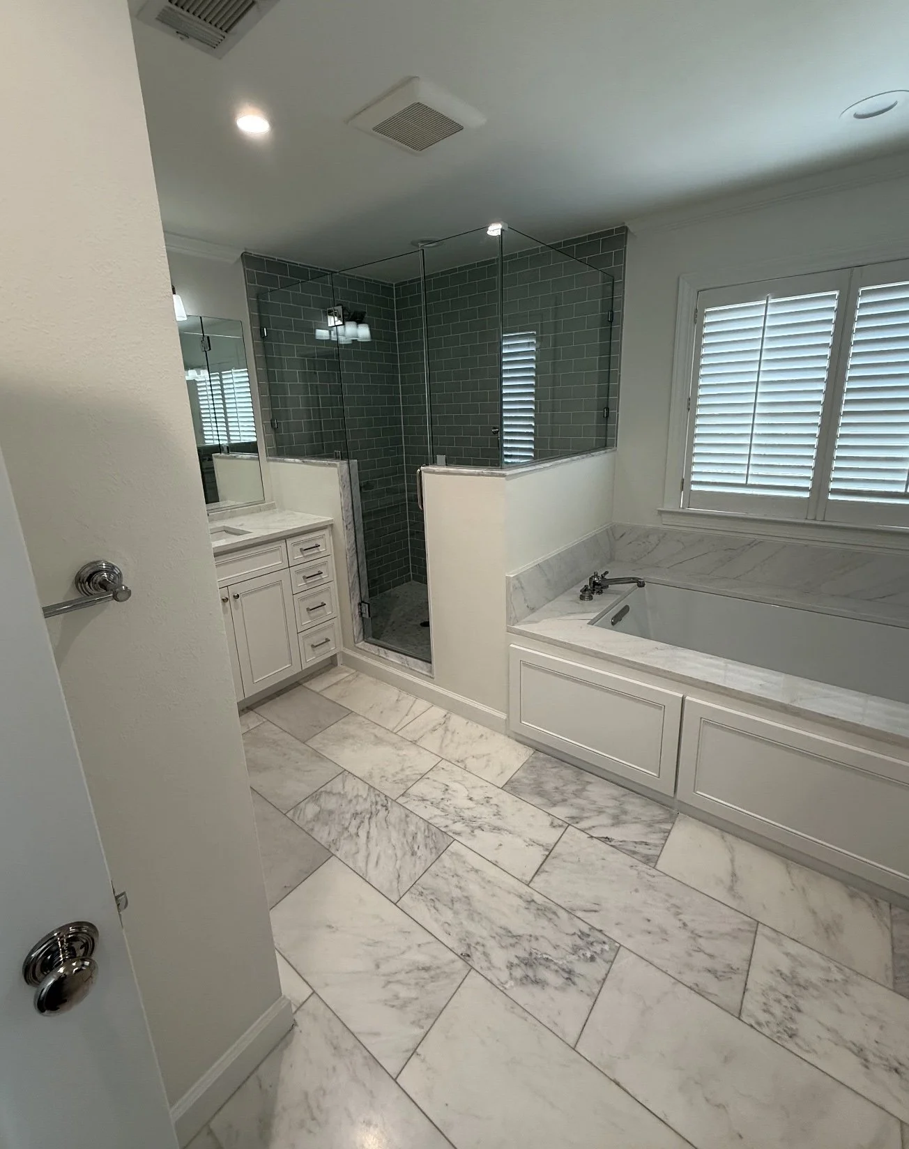 A luxurious bathroom with a marble floor, a glass-enclosed shower with dark green tile walls, a built-in bathtub with marble surround, and a vanity with storage drawers and a mirror. White plantation shutters cover a window.